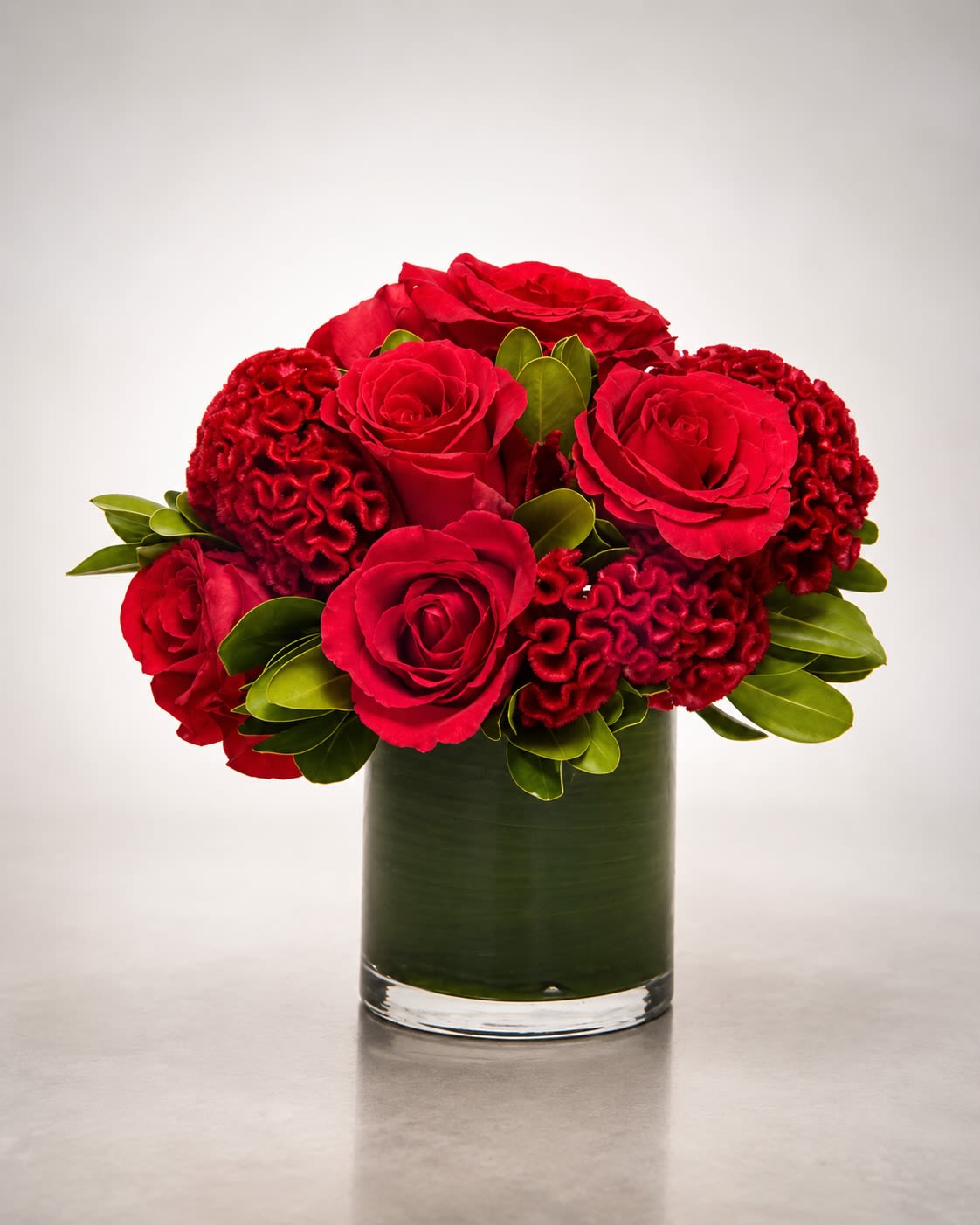 Velvet Rouge - A rich and romantic floral arrangement featuring premium red roses paired with dramatic red celosia blooms designed in a modern leaf-lined cylinder vase. Velvet Rouge combines classic romance with luxurious texture, creating a bold monochromatic presentation filled with depth, elegance, and contemporary style.  The velvety red roses are complemented by sculptural celosia blooms that add movement and unique texture throughout the arrangement, while polished greenery and tropical leaf lining provide a clean upscale finish. This arrangement is perfect for anniversaries, romantic gestures, Valentine’s Day, luxury gifting, or elegant modern décor with a bold statement feel.  Due to seasonal and market availability, certain flower varieties or foliage may occasionally vary while maintaining the arrangement’s rich crimson palette, fullness, and luxurious overall aesthetic. Vase style and foliage lining may also vary slightly depending on availability while preserving the same elevated presentation and contemporary design style.