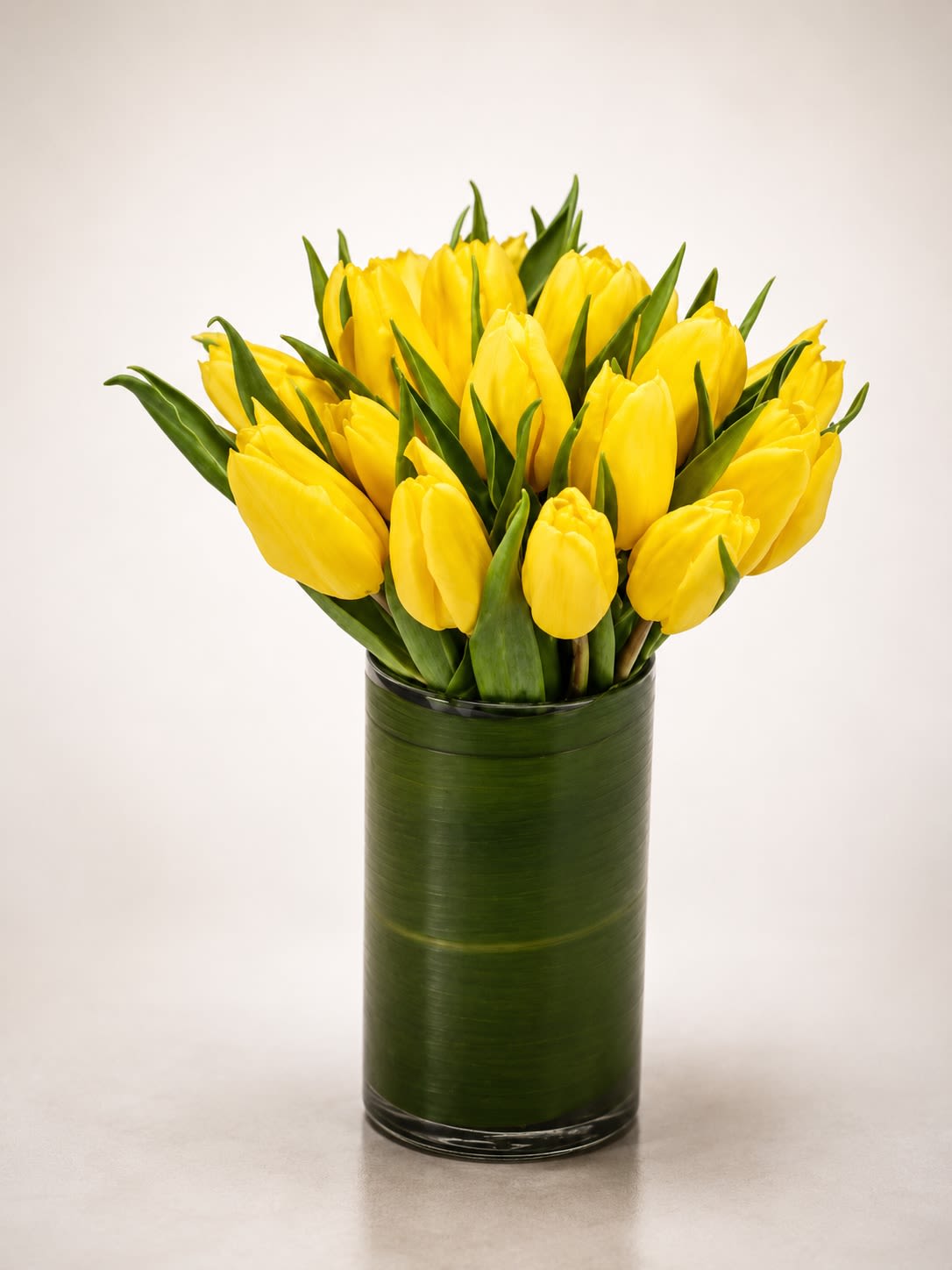 Golden Soleil Tulips - A radiant arrangement of vibrant yellow tulips artfully gathered in a sleek leaf-lined glass cylinder vase. Golden Soleil Tulips captures the warmth of sunshine and the elegance of modern floral design, making it a beautiful gift for birthdays, celebrations, thank-you gestures, get well wishes, or simply brightening someone’s day.  Designed with premium tulips in a clean contemporary style, this arrangement offers a fresh, cheerful, and sophisticated look perfect for homes, offices, and modern interiors. The rich green foliage lining the vase adds a polished luxury touch while allowing the bright golden blooms to stand out beautifully.  Due to seasonal and market availability, tulip shades may vary slightly from golden yellow to softer buttery tones. Vase style and foliage accents may also vary while maintaining the arrangement’s modern minimalist aesthetic and overall color palette.