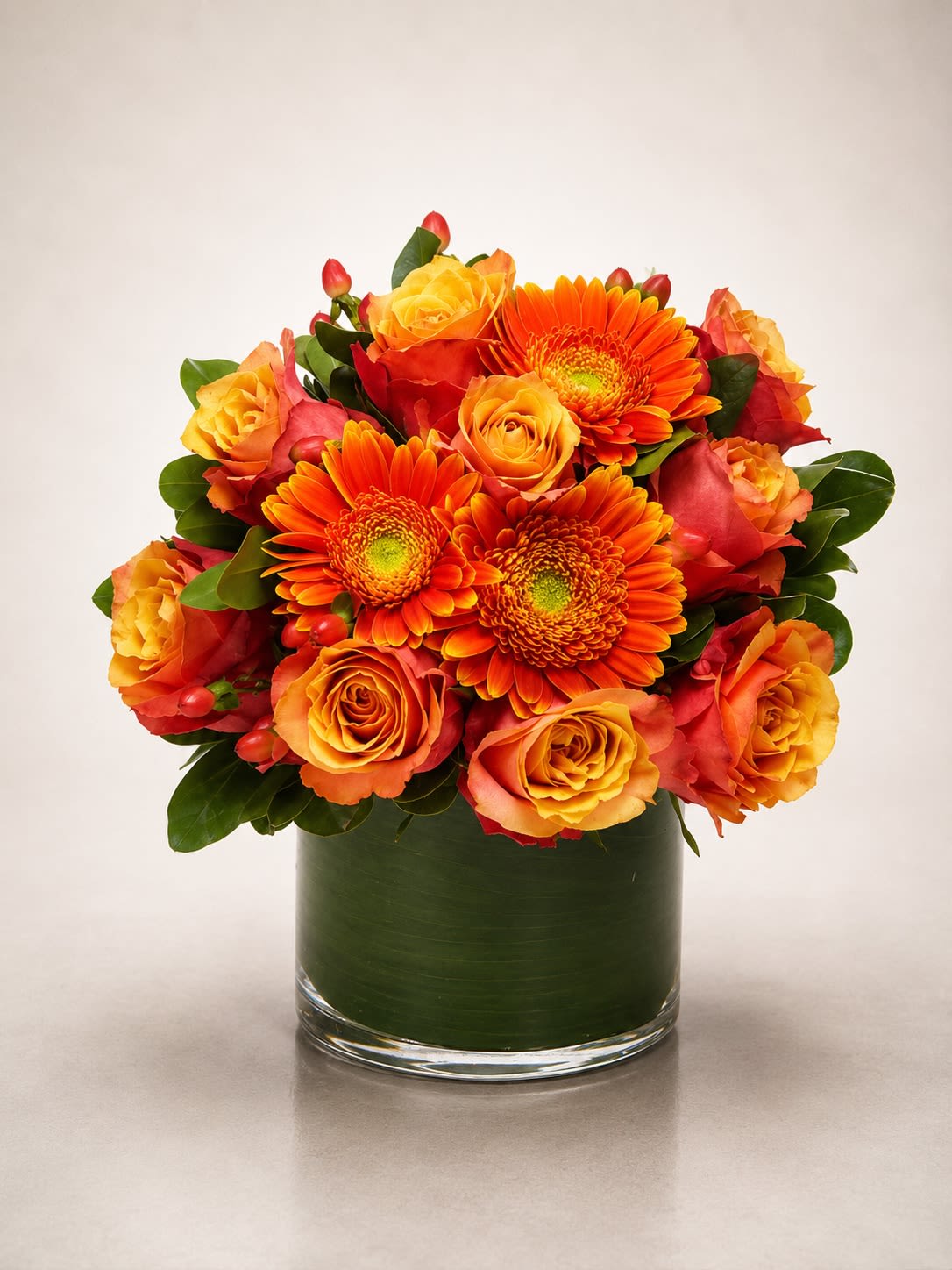 Citrus Sunset Bloom - A vibrant luxury arrangement featuring fiery orange gerbera daisies, golden sunset roses, and seasonal accent blooms artfully designed in a modern leaf-lined glass cylinder vase. Citrus Sunset Bloom radiates warmth, happiness, and bold contemporary elegance with its rich sunset-inspired palette of orange, gold, coral, and amber tones.  Designed with premium roses and vivid gerbera daisies accented by lush greenery and textural seasonal elements, this arrangement is perfect for birthdays, celebrations, thank-you gifts, fall-inspired décor, congratulations, or simply bringing energy and joy into any room. Its bold color story and compact modern design create a fresh, upscale floral statement.  Due to seasonal and market availability, flower varieties and accent blooms may occasionally vary while maintaining the arrangement’s warm sunset-inspired palette, luxurious fullness, and contemporary style. Vase style and foliage lining may also vary slightly while preserving the same elegant presentation.