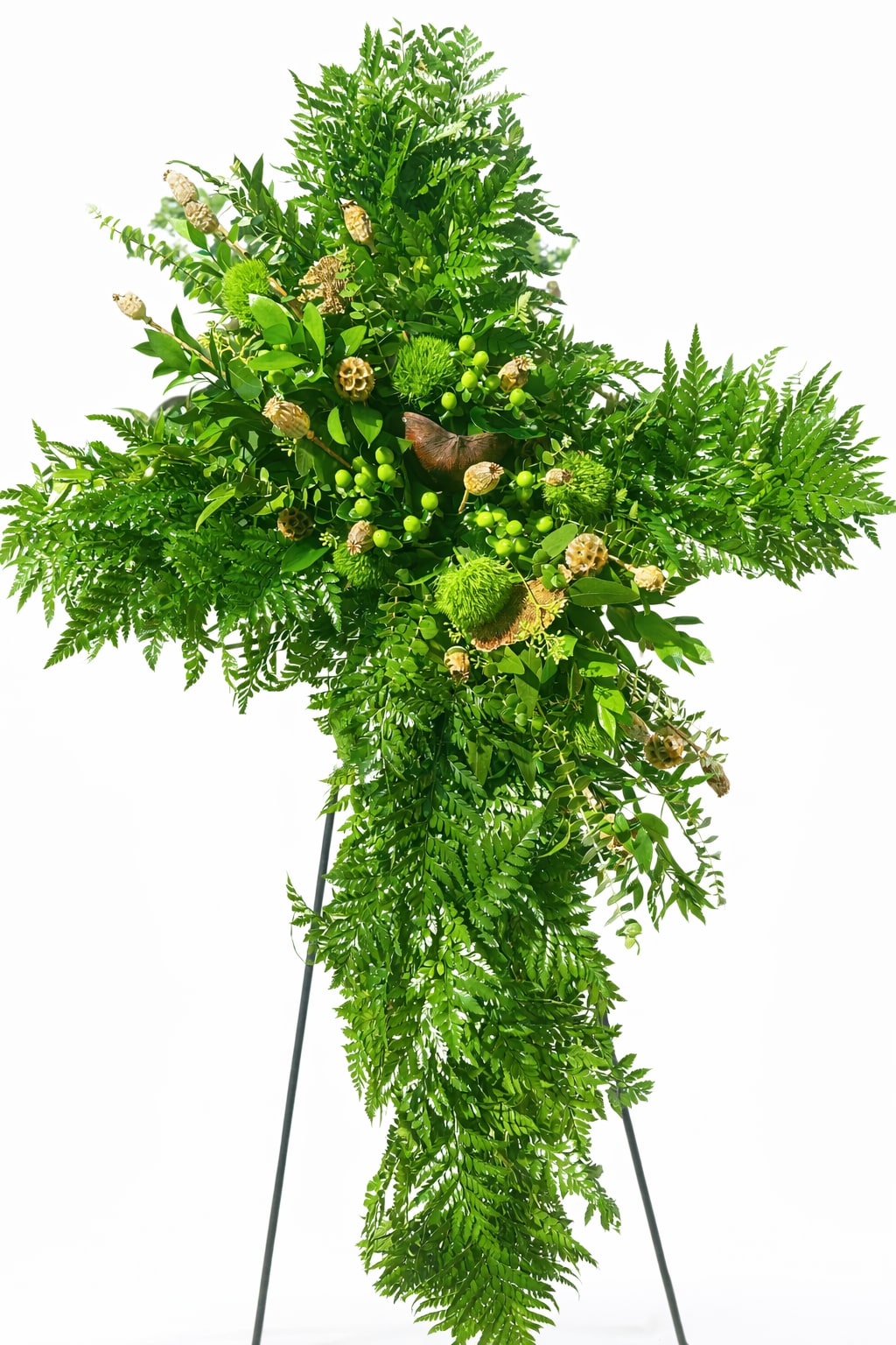 Natures Promise Tribute - A dignified standing cross formed entirely from lush green foliage creates a serene, reverent tribute perfect for a sympathy arrangement or memorial service. The vertical silhouette of this standing cross frame speaks to solemn respect and quiet remembrance, making it an appropriate choice for a funeral or graveside tribute when you want the person you're honoring to be remembered with grace.  Layered ferns and varied leafy branches build a full, textured cross with depth and movement; small pale seed pods and bright green berry-like accents are scattered through the center to give subtle contrast and visual interest while staying within a calming green palette. Mounted on a sturdy standing cross frame, this piece is sized for display at a service or beside a gravesite and conveys a gentle, natural presence without flowers, ideal for families seeking a classic, evergreen expression of sympathy.  Designed to comfort the bereaved, this tribute sends a message of peaceful remembrance to the family and loved ones of the deceased. Order with delivery timing for the service or cemetery, and our team will handle setup on the standing frame so the cross arrives ready to honor the memory in a respectful, lasting way.