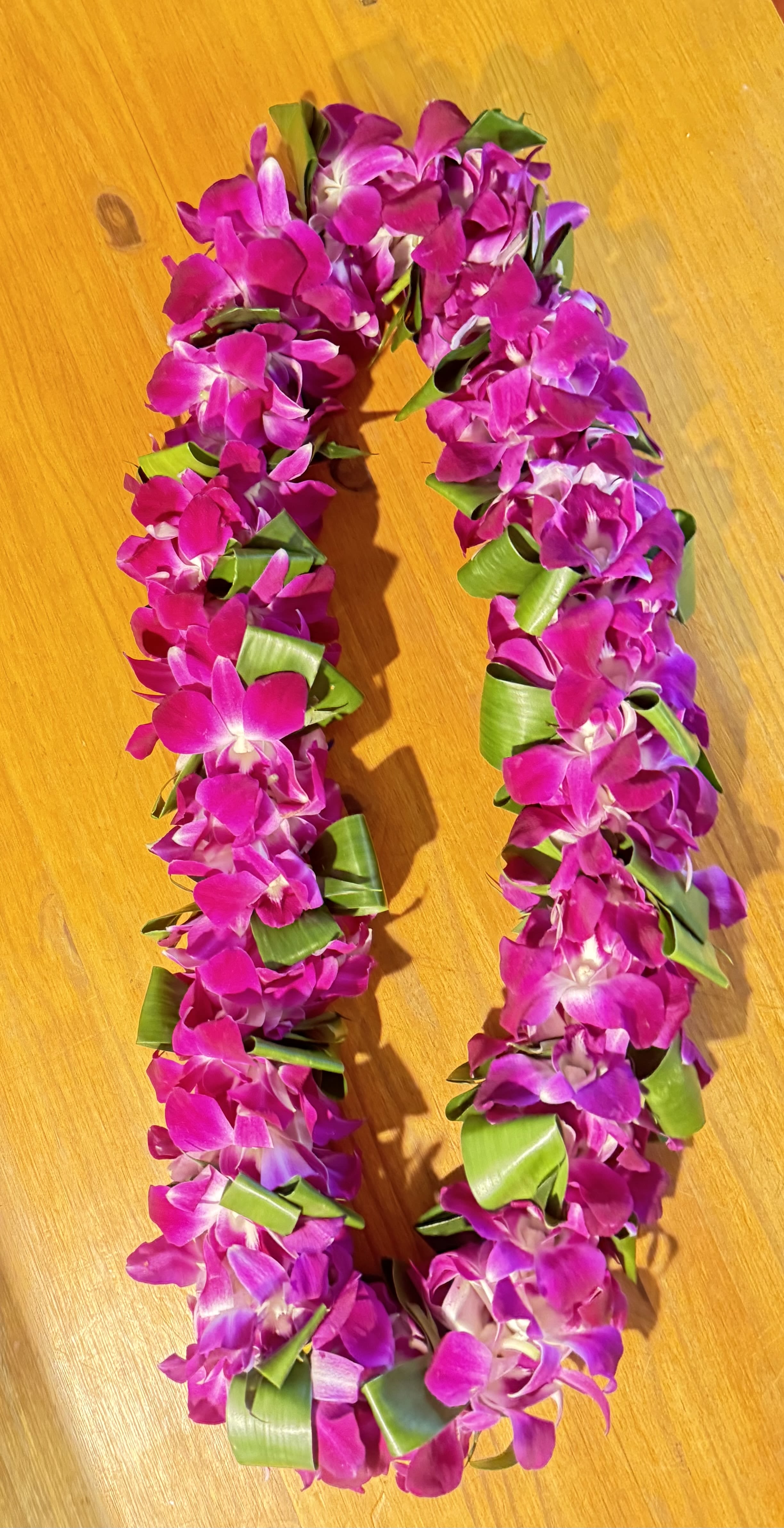 Bombai double orquid lei with tea leaves - Bright, densely strung  orchid lei with small white centers and folded green tea leaf accents-an island-inspired flower arrangement perfect as a graduation lei, prom accessory, or summer celebration adornment. The compact circular form sits close to the neck for a festive, celebratory look that makes the recipient feel honored and seen.  Handcrafted from tropical orchids in vibrant  with crisp white centers and glossy green tea leaves folded into the design, this lei is densely packed for a lush, saturated appearance. The compact size and sturdy construction keep blooms secure during processions and photos; treat gently, keep cool, and mist lightly to extend freshness. Ideal for marking milestone moments or gifting to someone you want to celebrate in bright, tropical style.