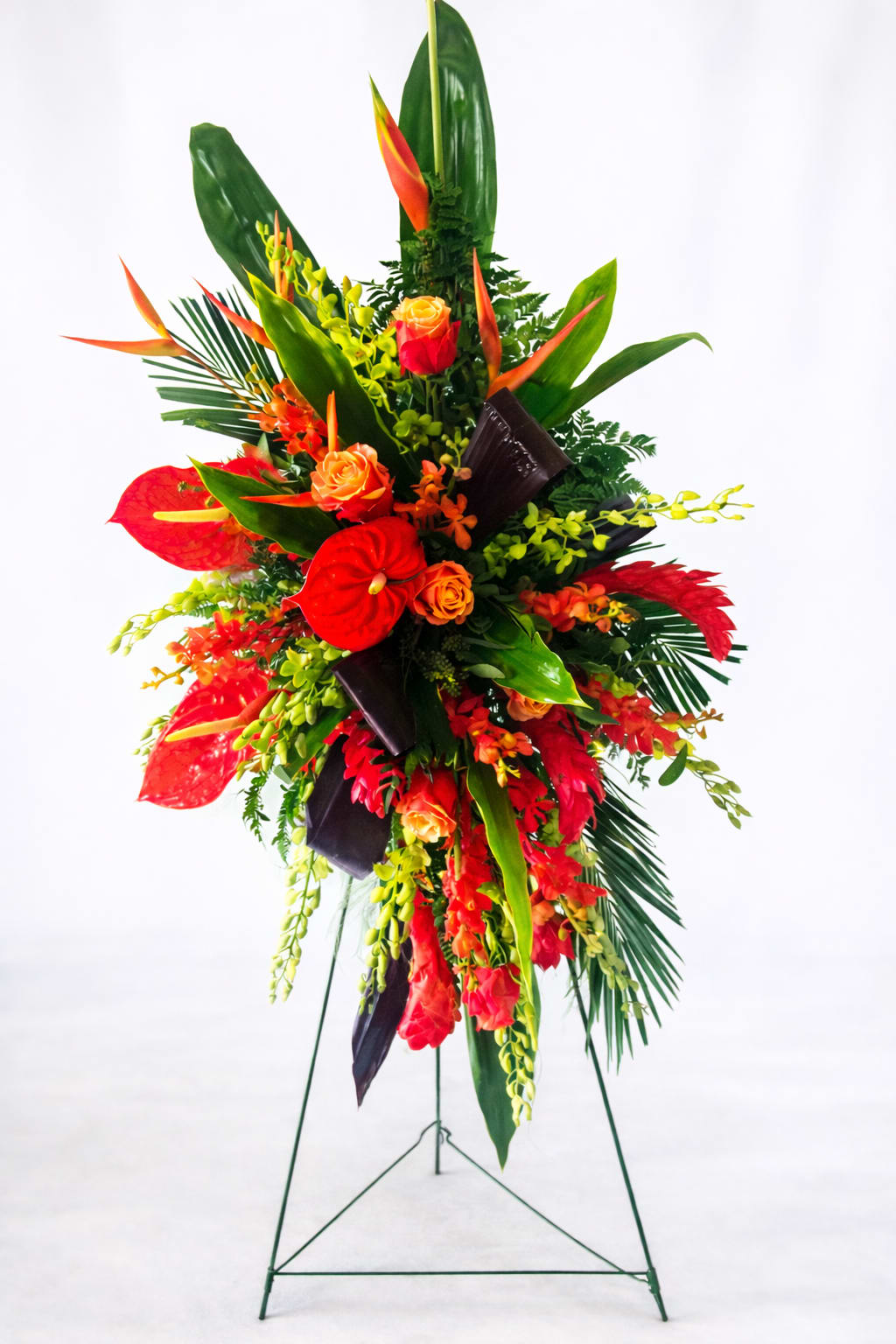 Paradise Remembrance - A dramatic, tall tropical standing spray designed for sympathy and memorial services, this floral arrangement makes a bold, respectful statement. The vertical silhouette and dense, layered composition create an immediate sense of presence-perfect for honoring someone with a formal display at a funeral or memorial gathering, or as a striking piece for a large event.  Bright red and orange roses and tropical blooms are paired with glossy anthuriums and sprays of yellow orchids, set against long, dark green leaves and accent foliage for high-contrast impact. The colors read warm and vivid from a distance while offering rich texture up close: velvety rose petals, sculptural anthurium hearts, and delicate orchid clusters combine to create a balanced, ceremonial look.  Arranged on a sturdy metal easel, this standing spray is built to command attention and convey heartfelt respect to the person you're honoring. Sized for display at services or in a reception space, it arrives ready to stand; minimal care is needed-keep away from direct heat and mist foliage lightly if necessary to maintain freshness.