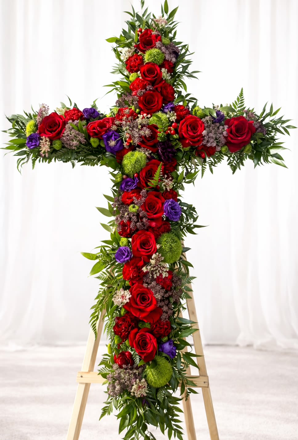Royal Remembrance  - A striking cross of densely packed red roses makes an immediate, reverent impression-perfect as a sympathy arrangement or memorial tribute to honor a loved one. This funeral cross arrangement presents a formal, solemn presence ideal for a memorial service or graveside display, offering a dignified way to express sympathy and respect to the family you're thinking of.  The design pairs deep red roses with purple accents, bright green pom-pom chrysanthemums, white filler flowers, and abundant foliage that softens the geometric silhouette. The arrangement is built upright on a wooden easel stand so it reads clearly from a distance; the tall, symmetrical shape creates a powerful visual focal point at services and viewings.  Made to convey heartfelt remembrance, this piece is suited for funerals, memorial ceremonies, or as a standing tribute delivered to a service. The wooden easel ensures stable display-please let us know service time and location when ordering so we can coordinate delivery and placement for the moment that matters.