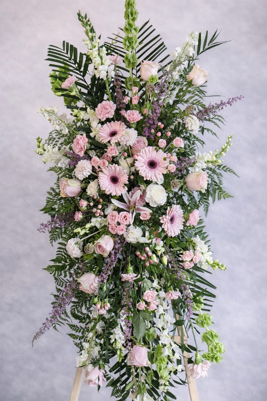 Softly Remembered - A gentle vertical spray of pastel blooms, this sympathy flower arrangement is designed to comfort and honor at a memorial service. The tall standing spray for sympathy places soft pink gerbera daisies and pale roses at the visual center, surrounded by white lilies and carnations to create a calm, reverent presence that reads as both formal and tender.  Textured accents of snapdragons, stock, and spray roses weave through abundant green foliage, forming a dense, airy silhouette on a wooden easel that gives the tribute height and dignity. The palette-pink, white, green, and touches of purple-keeps the focus soothing and respectful, while the mix of large focal flowers and smaller clustered blooms provides visual depth and gentle movement.  Perfect for expressing heartfelt condolences to family or honoring the memory of a loved one, this arrangement conveys sympathy without overwhelming. Sized to stand prominently at a service or graveside, it arrives ready on its easel; minimal care is needed beyond keeping it cool and sheltered from direct sun while on display.