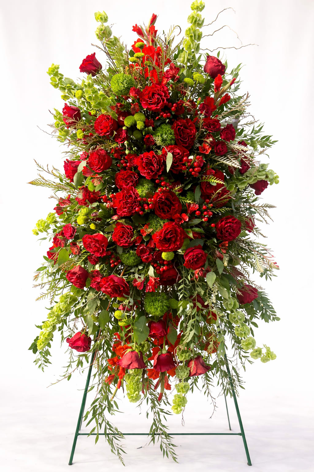 Strength of Love Tribute - Vivid red roses and clustered red carnations form a dramatic, upright tribute that reads with quiet strength-perfect as a sympathy standing spray or memorial arrangement to honor a loved one. The predominance of red paired with bright green pompon chrysanthemums and pale green hanging accents creates a bold, respectful statement that feels both formal and heartfelt for a funeral or remembrance service.  This large standing spray is densely composed for substantial height and fullness, mounted on a metal easel for prominent display. Textural contrast comes from the mix of roses, carnations, chrysanthemums and airy foliage that extends outward and downward, giving the piece an elegant, sculpted silhouette in red, green, and white tones.  Choose this arrangement when you want the person you're remembering to feel deeply honored-the kind of floral tribute that conveys enduring affection and respect. The display arrives ready on its easel for immediate placement at a service; no special care is required beyond keeping the flowers in a cool location until the service begins.