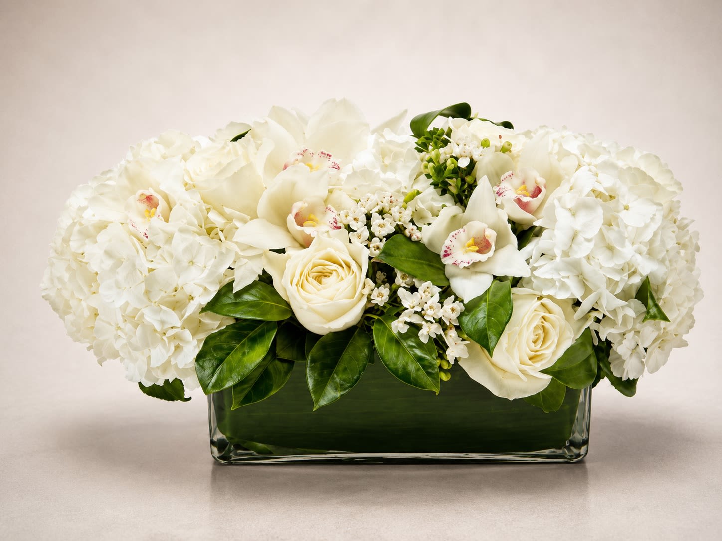 Ivory Garden Luxe - A luxurious all-white floral arrangement featuring lush hydrangeas, elegant white roses, delicate cymbidium orchids, and soft seasonal accent blooms artfully designed in a sleek modern glass rectangle vase. Ivory Garden Luxe embodies timeless sophistication with its serene monochromatic palette, abundant texture, and upscale contemporary styling.  Designed with premium blooms in creamy ivory and crisp white tones, this arrangement creates a refined statement perfect for sympathy gestures, weddings, anniversaries, luxury gifting, corporate settings, or elegant home décor. The combination of hydrangeas, orchids, roses, and delicate floral accents gives this piece a romantic garden-inspired feel while maintaining a polished modern aesthetic.  Due to seasonal and market availability, certain flower varieties, orchid accents, or greenery may occasionally vary while preserving the arrangement’s luxurious white palette, lush fullness, and elegant contemporary presentation. Vase style may also vary slightly depending on availability while maintaining the same upscale design.
