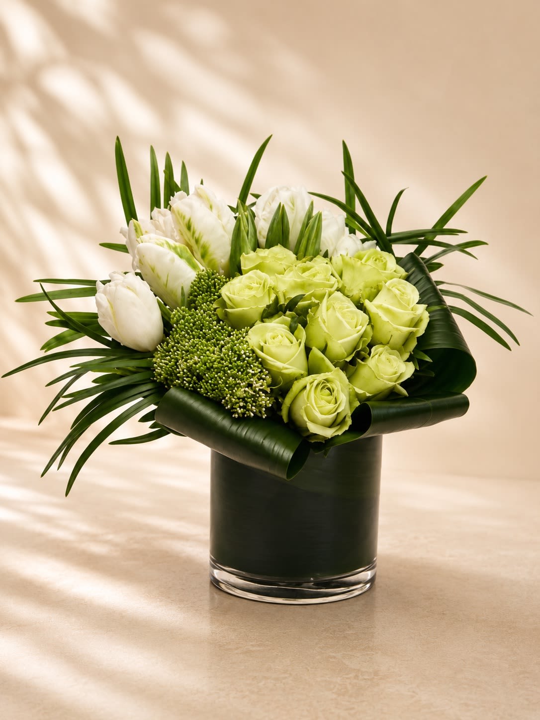 Sage Garden Luxe - A refined modern arrangement featuring soft green roses, crisp white tulips, elegant white blooms, and lush textural greenery artfully designed with sculptural tropical foliage in a sleek contemporary vase. Sage Garden Luxe blends fresh botanical tones with sophisticated floral styling to create a serene and upscale designer arrangement.  The calming palette of soft sage greens and creamy whites symbolizes renewal, peace, elegance, and prosperity, making this arrangement ideal for sympathy occasions, corporate gifting, birthdays, thank-you gestures, housewarmings, or sophisticated everyday décor. Its modern garden-inspired composition delivers a luxurious yet organic aesthetic perfect for contemporary interiors.  Due to seasonal and market availability, specific flower varieties or greenery accents may occasionally vary while maintaining the arrangement’s elegant green-and-white palette and modern artistic design. Vase style may also vary slightly depending on availability while preserving the same upscale presentation.