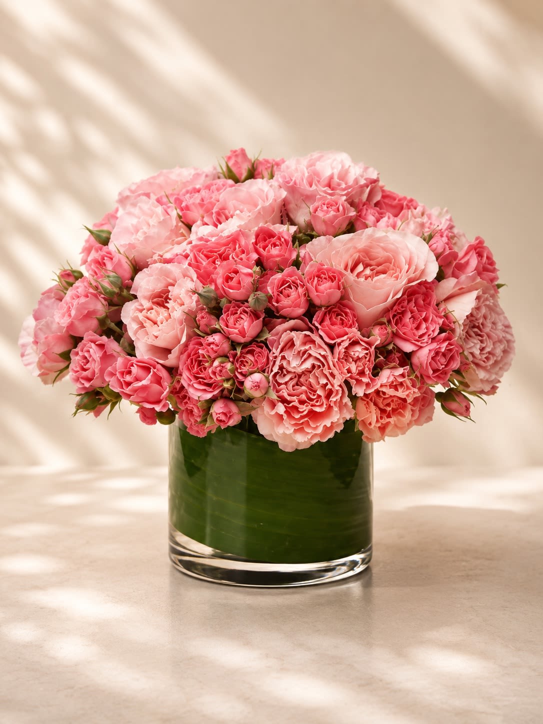 Blush Petal Romance - A lush and romantic arrangement overflowing with soft pink garden roses, delicate spray roses, and ruffled premium carnations artfully gathered in a modern leaf-lined glass vase. Blush Petal Romance captures feminine elegance and timeless beauty through layers of textured pink blooms in dreamy blush and rose tones.  This luxurious floral design symbolizes admiration, sweetness, grace, and affection, making it a perfect choice for birthdays, anniversaries, baby showers, romantic gestures, Mother’s Day, or elegant everyday gifting. The abundance of layered petals and monochromatic pink palette create a rich, couture-inspired arrangement with a soft luxurious feel.  Due to seasonal and market availability, specific rose or carnation varieties may vary slightly while maintaining the arrangement’s signature blush pink palette, fullness, and romantic aesthetic. Vase style may occasionally vary depending on availability while preserving the same upscale presentation.