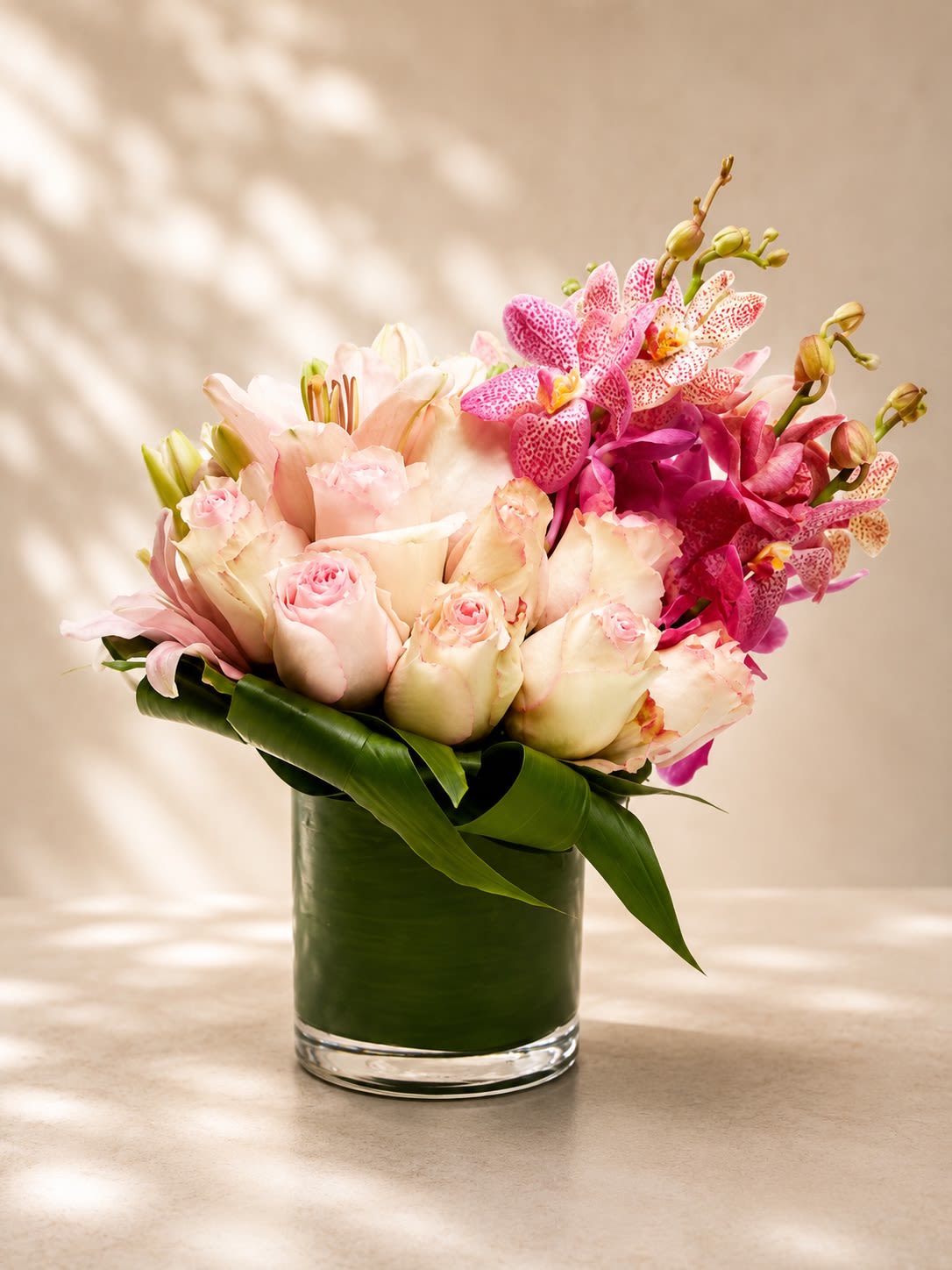 Orchid Blush Couture - A breathtaking luxury arrangement featuring blush pink roses, elegant lilies, and exotic vibrant orchids artfully designed with sculptural tropical foliage in a modern leaf-lined glass vase. Orchid Blush Couture blends soft romantic tones with striking orchid accents to create a floral design that feels graceful, feminine, and undeniably luxurious.  This couture-inspired arrangement symbolizes beauty, admiration, elegance, and sophistication, making it an exceptional choice for birthdays, anniversaries, romantic celebrations, upscale gifting, Mother’s Day, or luxurious home décor. The combination of delicate blush roses and vibrant orchid blooms creates a rich layered composition with a modern tropical elegance.  Due to seasonal and market availability, specific orchid varieties, lily blooms, or rose shades may vary slightly while maintaining the arrangement’s signature blush and pink palette and luxurious aesthetic. Vase style may occasionally vary depending on availability while preserving the same upscale presentation.