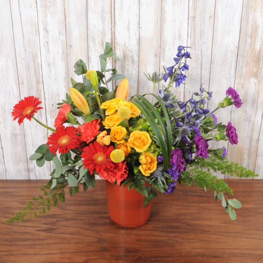 Bright mixed arrangement with red gerberas, yellow roses, lilies, and purple blooms in an orange vase