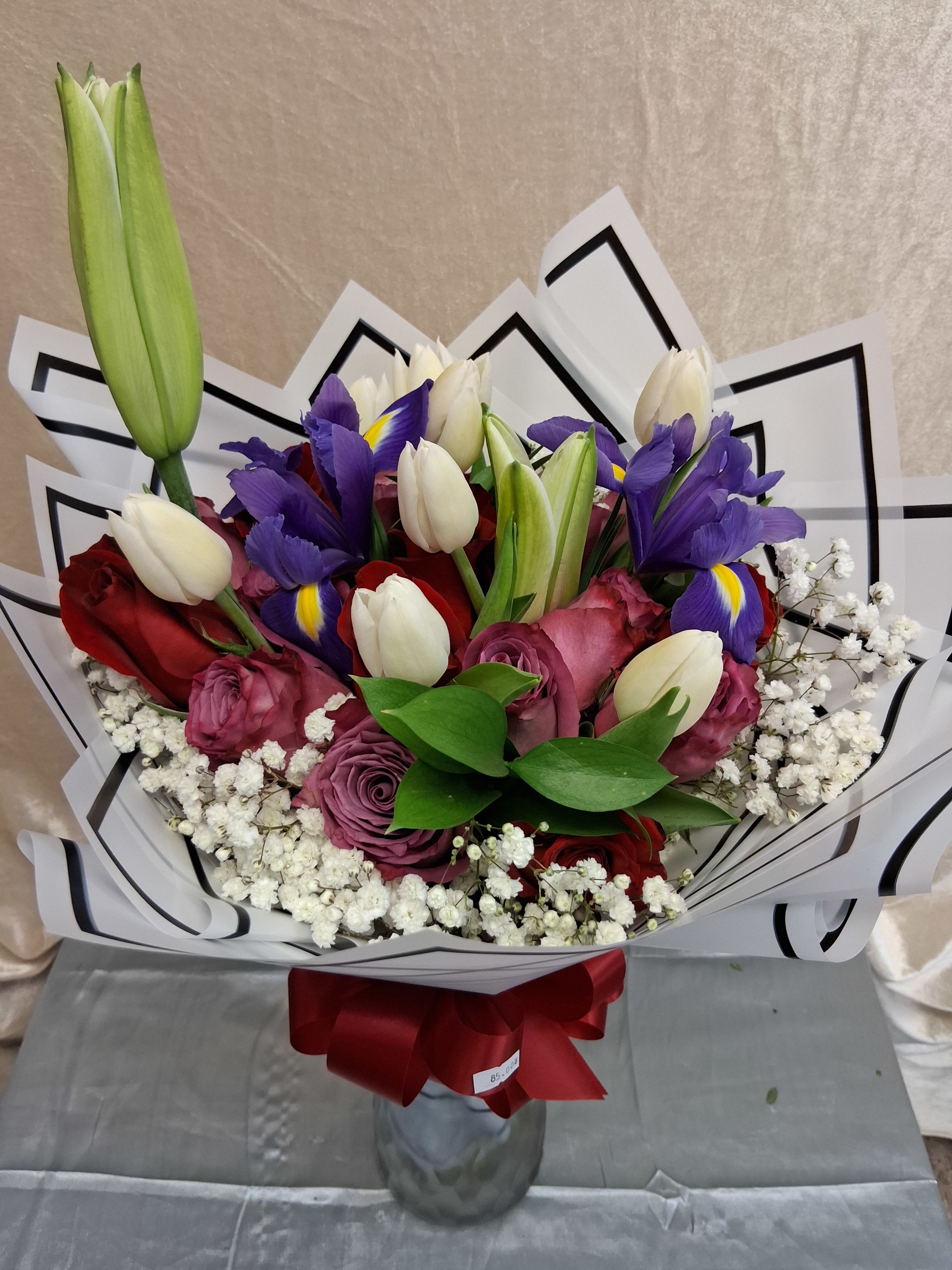 2 dozen roses purple and red with lily's and baby breath bouquet  - Deep, romantic color takes center stage in this flower arrangement-perfect as an anniversary bouquet or a love-themed gift. Two dozen roses mingle with purple irises and white tulips to create a lush, rounded bouquet that reads both classic and contemporary; the combination of red, purple, white, and touches of pink makes a striking impression that says "I cherish you" without words.  The arrangement is finished in a clear glass vase and wrapped in black-and-white geometric paper with a red ribbon tied at the base for a polished, gift-ready look. White baby's breath softens the stronger hues along the lower edge, giving the bouquet a layered, full shape and delicate texture. Sized to feel substantial, it's arranged to display varied heights-roses and irises stand out while tulips and baby's breath add lightness and movement.  Give this to the person you want to make feel deeply celebrated-ideal for anniversaries, romantic occasions, or a wedding-related presentation. To enjoy it longer, place the vase in a cool spot away from direct sunlight and change the water every other day; if you have any special delivery requests, include them when you order.