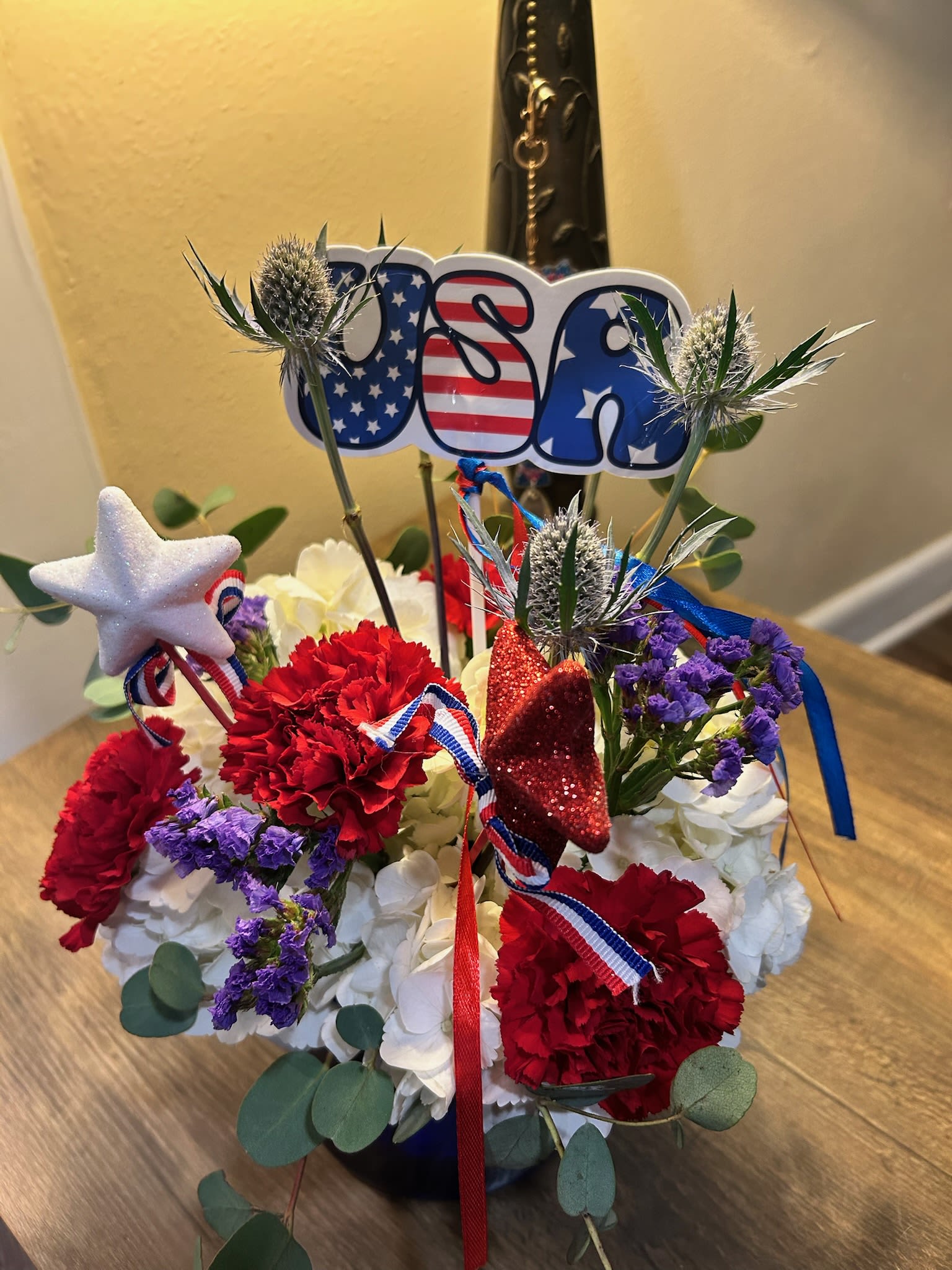 Happy Birthday USA - Bright red carnations, snowy white hydrangeas, and pops of purple filler blooms combine in a compact, rounded birthday arrangement that reads festive and proudly patriotic-perfect as a birthday bouquet or Fourth of July surprise. The USA topper and glittery star picks lift the design with playful height while red, white, and blue ribbon accents reinforce the celebratory mood; the person you're celebrating will feel cheered and noticed the moment this arrives.  This arrangement sits in a clear vase and is finished with eucalyptus that spills around the base, softening the bold color blocks with a casual, hand-crafted texture. The mix of carnations and hydrangeas creates a lush, long-lasting display with sturdy stems and generous volume; star decorations and ribbon add sparkle and movement without overwhelming the flowers.  Give this to a patriotic friend, a family member marking a summer birthday, or anyone who loves bold, colorful florals that read both festive and thoughtful. Arrange for delivery on the special day and include a short note-the sturdy flowers and vase make it easy to display, and simple care (fresh water and a cool spot away from direct sun) will help the bouquet stay lively for days.