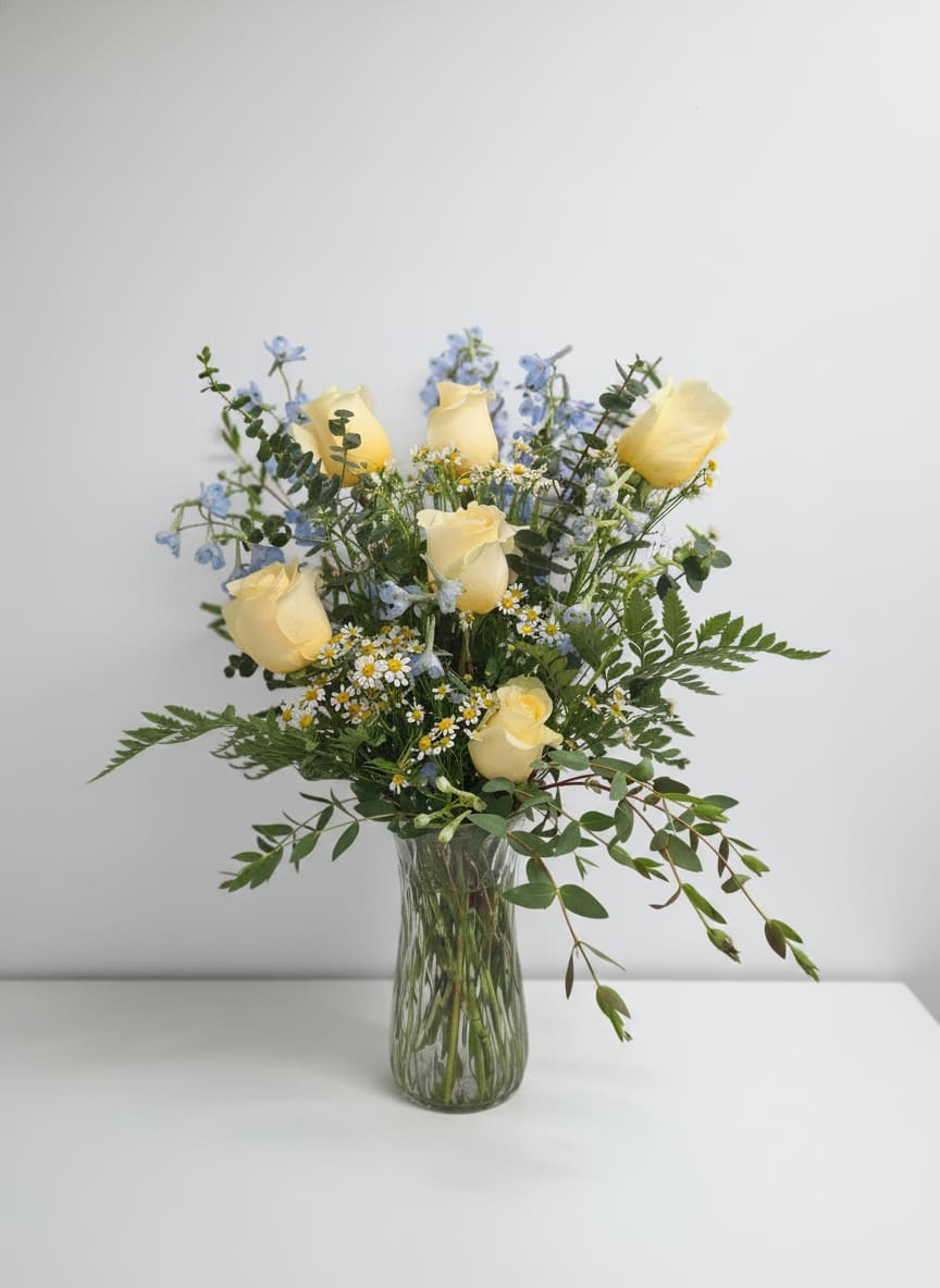 Country Blues - Pale yellow roses and airy blue delphinium come together in a relaxed garden-style bouquet that feels like a sunny morning-perfect as a birthday bouquet, a thank-you arrangement, or a cheerful spring gift. The combination of soft pastels and fresh greenery creates a calm, joyful impression so the person you're thinking of will feel genuinely seen and brightened.  Clusters of small white-and-yellow daisy-like blooms add playful texture against the fuller rose heads, while stems and foliage extend naturally from a clear glass vase for an unfussy, hand-gathered look. The palette of yellow, blue, white, and green keeps the arrangement versatile and seasonally fresh; dimensions follow a loose, airy silhouette that fills a tabletop without overwhelming it.  Send this when you want to lift someone's spirits, celebrate a small win, or say thanks in a way that feels personal and light. The arrangement arrives in a clear glass vase ready to display-simple fresh-water care (trim stems every few days, change water regularly) will keep it looking its best.