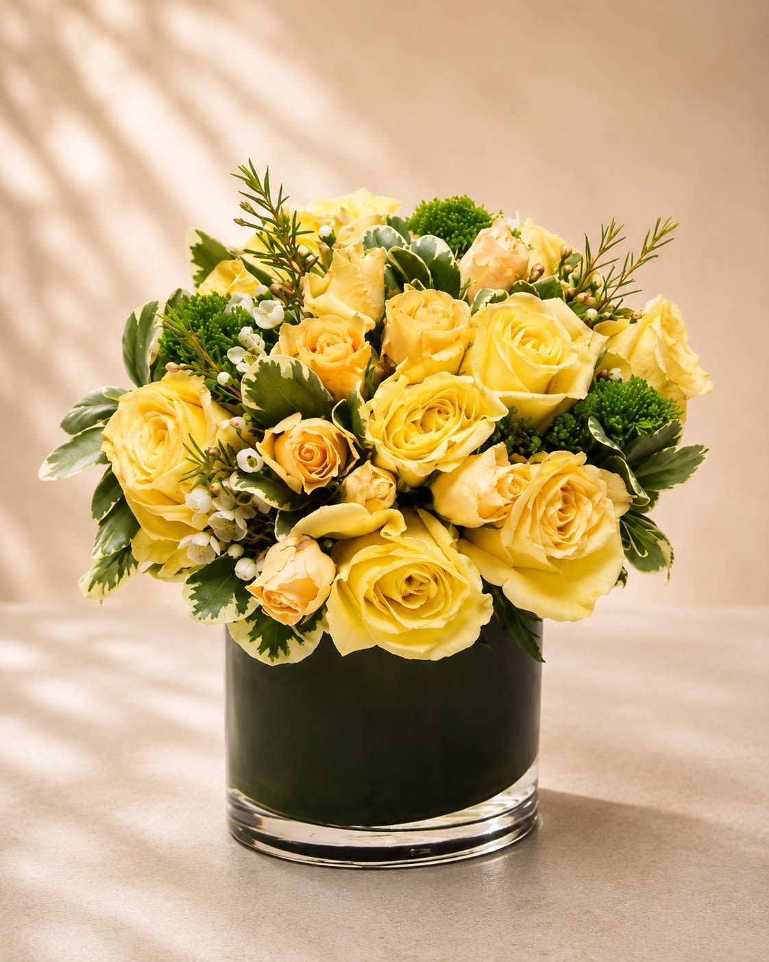 Golden Meadow Luxe - A radiant and elegant floral arrangement featuring buttery yellow roses accented with fresh green textures, delicate wax flowers, and lush variegated foliage arranged in a modern leaf-lined glass vase. Golden Meadow Luxe blends cheerful warmth with refined sophistication, creating a design that feels both uplifting and luxuriously garden-inspired.  Yellow roses symbolize friendship, joy, happiness, and new beginnings, making this arrangement a beautiful choice for birthdays, congratulations, thank-you gifts, get well wishes, celebrations, or simply brightening someone’s day. The soft golden tones paired with crisp greenery create a fresh and timeless aesthetic perfect for both modern and classic spaces.  Due to seasonal and market availability, greenery varieties and filler flowers may vary slightly while preserving the arrangement’s signature golden-yellow palette and elegant garden style. Vase style may occasionally vary depending on availability while maintaining the same premium presentation.