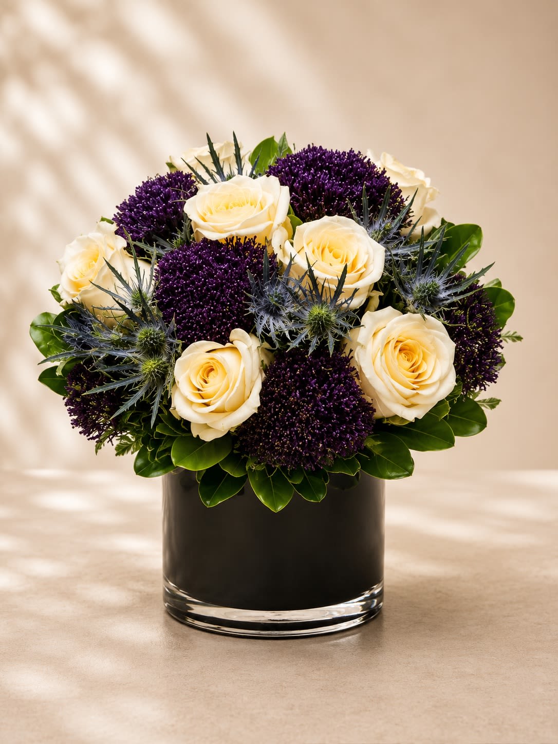 Midnight Amethyst Luxe - A bold and sophisticated floral arrangement featuring creamy ivory roses paired with dramatic deep-purple allium blooms, textured blue thistle accents, and rich glossy greenery arranged in a sleek black leaf-lined glass vase. Midnight Amethyst Luxe is designed with modern elegance and artistic contrast, blending soft romance with striking contemporary textures for a truly couture-inspired presentation.  The ivory roses symbolize grace, admiration, and timeless beauty, while the rich amethyst-purple florals add mystery, luxury, and depth. The sculptural blue thistle accents create an elevated botanical feel, making this arrangement perfect for upscale gifting, modern home décor, birthdays, anniversaries, corporate events, or anyone who appreciates unique and luxurious floral artistry.  Due to seasonal and market availability, specialty blooms and textured accents may vary slightly while preserving the arrangement’s signature ivory, deep purple, and cool-toned palette and modern luxury aesthetic. Vase style may occasionally vary depending on availability while maintaining the same premium presentation.