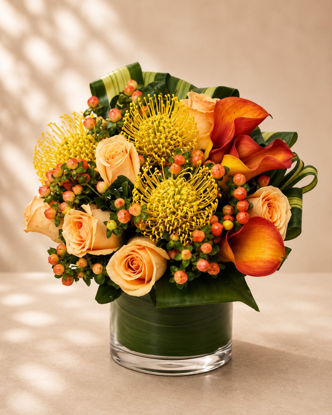 Sunset Safari Couture - An exotic and artfully designed floral arrangement featuring warm peach roses, fiery sunset calla lilies, golden pincushion protea, vibrant hypericum berries, and lush tropical foliage arranged in a modern leaf-lined glass vase. Sunset Safari Couture blends bold texture, rich color, and contemporary floral artistry to create a striking luxury arrangement inspired by tropical sunsets and high-end botanical design.  The golden protea symbolizes strength, transformation, and uniqueness, while the warm peach and amber tones evoke warmth, celebration, and sophistication. This arrangement is perfect for upscale gifting, birthdays, corporate events, modern home décor, congratulations, or anyone who loves dramatic and exotic floral designs with a couture aesthetic.  Due to seasonal and market availability, specialty blooms such as protea or calla lilies may vary slightly while preserving the arrangement’s signature warm sunset palette and tropical luxury style. Vase style may occasionally vary depending on availability while maintaining the same premium presentation.