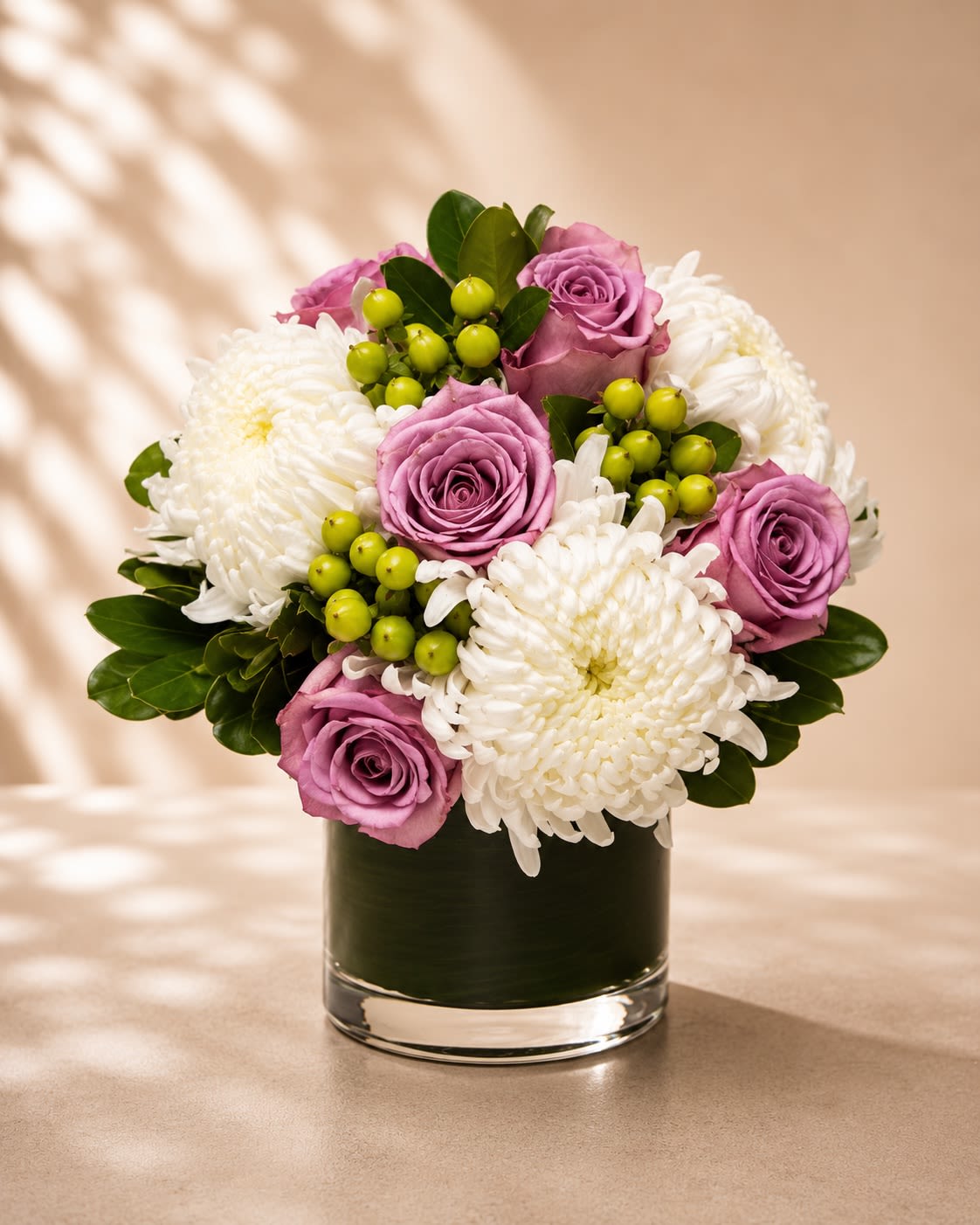 Lavender Whisper Luxe - A soft and elegant floral arrangement featuring lavender roses, lush white spider chrysanthemums, vibrant green hypericum berries, and rich glossy greenery arranged in a modern leaf-lined glass vase. Lavender Whisper Luxe blends delicate romance with contemporary sophistication, creating a refined design full of texture, serenity, and timeless beauty.  Lavender roses symbolize enchantment, grace, admiration, and elegance, while the crisp white blooms add purity and sophistication. The fresh green accents provide a modern botanical touch that enhances the arrangement’s luxurious and calming aesthetic. This arrangement is perfect for birthdays, sympathy gestures, thank-you gifts, anniversaries, wellness occasions, or elegant everyday décor.  Due to seasonal and market availability, floral varieties and greenery textures may vary slightly while preserving the arrangement’s signature lavender, white, and fresh green palette and luxurious garden-inspired style. Vase style may occasionally vary depending on availability while maintaining the same premium presentation.