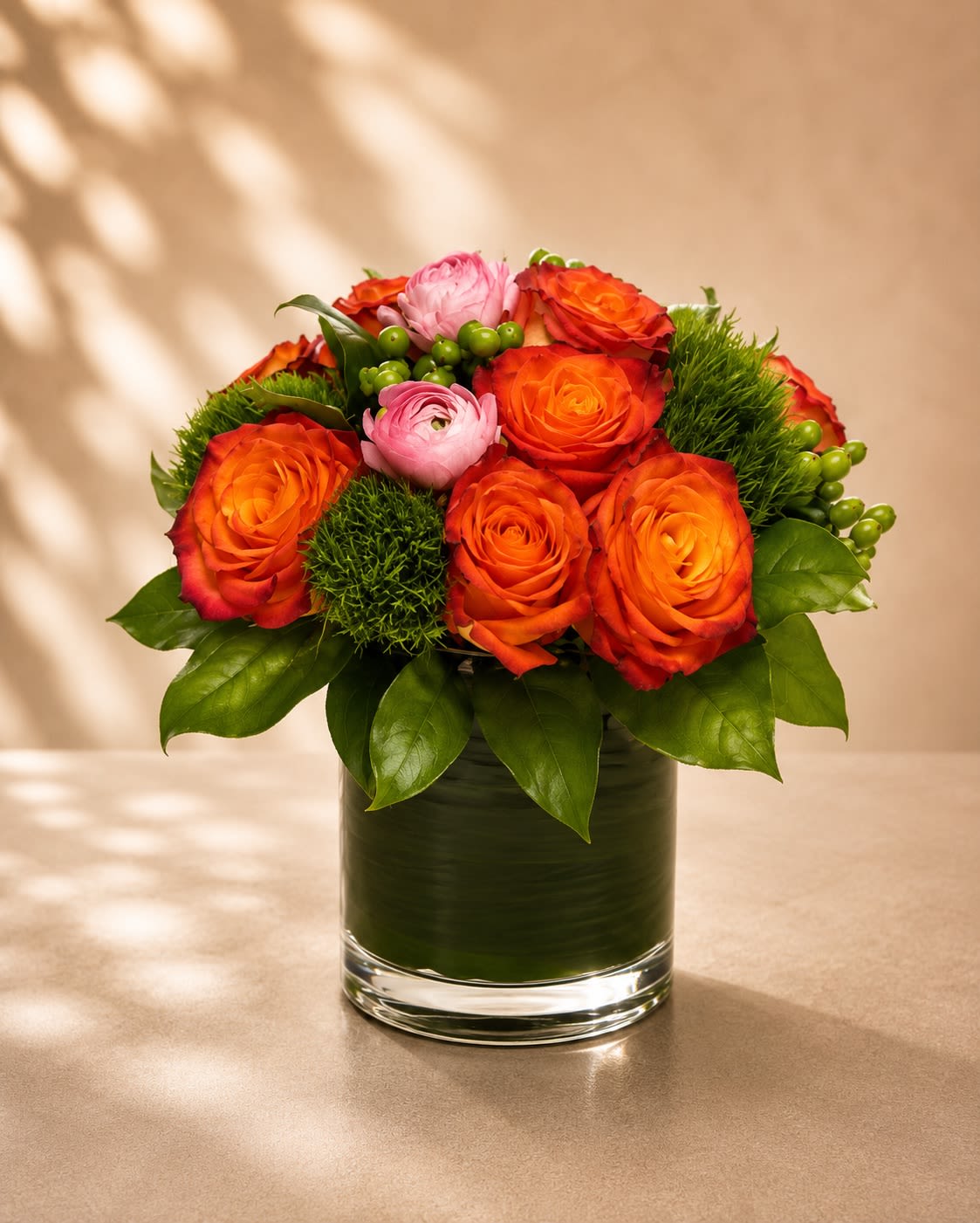 Ember Blush Garden - A vibrant designer arrangement featuring fiery orange roses, soft blush ranunculus, textured green dianthus, and fresh hypericum berries arranged in a modern leaf-lined glass vase. Ember Blush Garden blends warm sunset tones with soft romantic accents for a floral design that feels elegant, artistic, and full of life.  Orange roses symbolize passion, energy, and admiration, while blush ranunculus represents charm and radiant beauty. The textured green dianthus and bright berries add a playful modern touch, creating depth and movement throughout the arrangement. This luxurious design is perfect for birthdays, celebrations, thank-you gifts, anniversaries, or anyone who loves bold color with a soft romantic twist.  Due to seasonal and market availability, accent flowers and greenery may vary slightly while maintaining the arrangement’s signature vibrant palette and upscale modern style. Vase style may occasionally vary depending on availability while preserving the same elegant presentation.