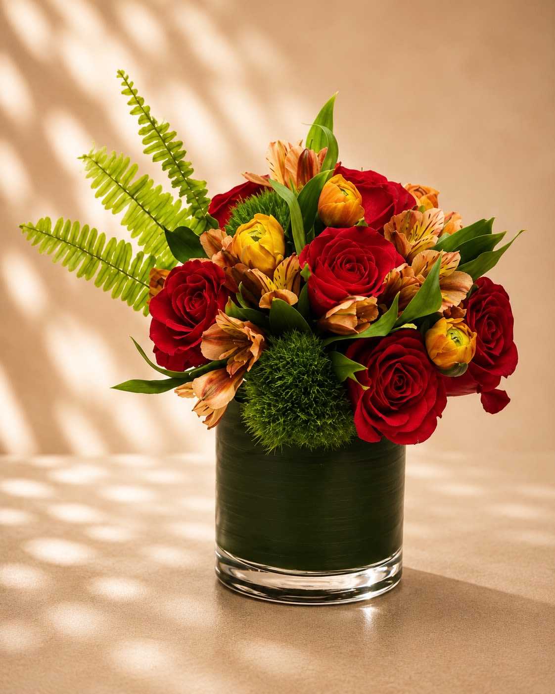 Crimson Ember Elegance - A rich and dramatic floral arrangement featuring luxurious red roses, golden ranunculus, warm bronze alstroemeria, textured green dianthus, and sculptural tropical greenery in a sleek leaf-lined glass vase. Crimson Ember Elegance combines deep romantic reds with glowing autumn-inspired accents for a floral design that feels bold, sophisticated, and modern.  Red roses symbolize deep love, passion, and timeless elegance, while golden ranunculus adds warmth, charm, and refined texture. The bronze alstroemeria and lush tropical foliage create depth and movement, giving this arrangement a contemporary designer feel with a romantic edge. Perfect for anniversaries, romantic gestures, birthdays, celebrations, or upscale gifting occasions.  Due to seasonal and market availability, accent flowers and greenery may vary slightly while maintaining the arrangement’s signature rich jewel-toned palette and luxurious modern style. Vase style may occasionally vary depending on availability while preserving the same elegant presentation.