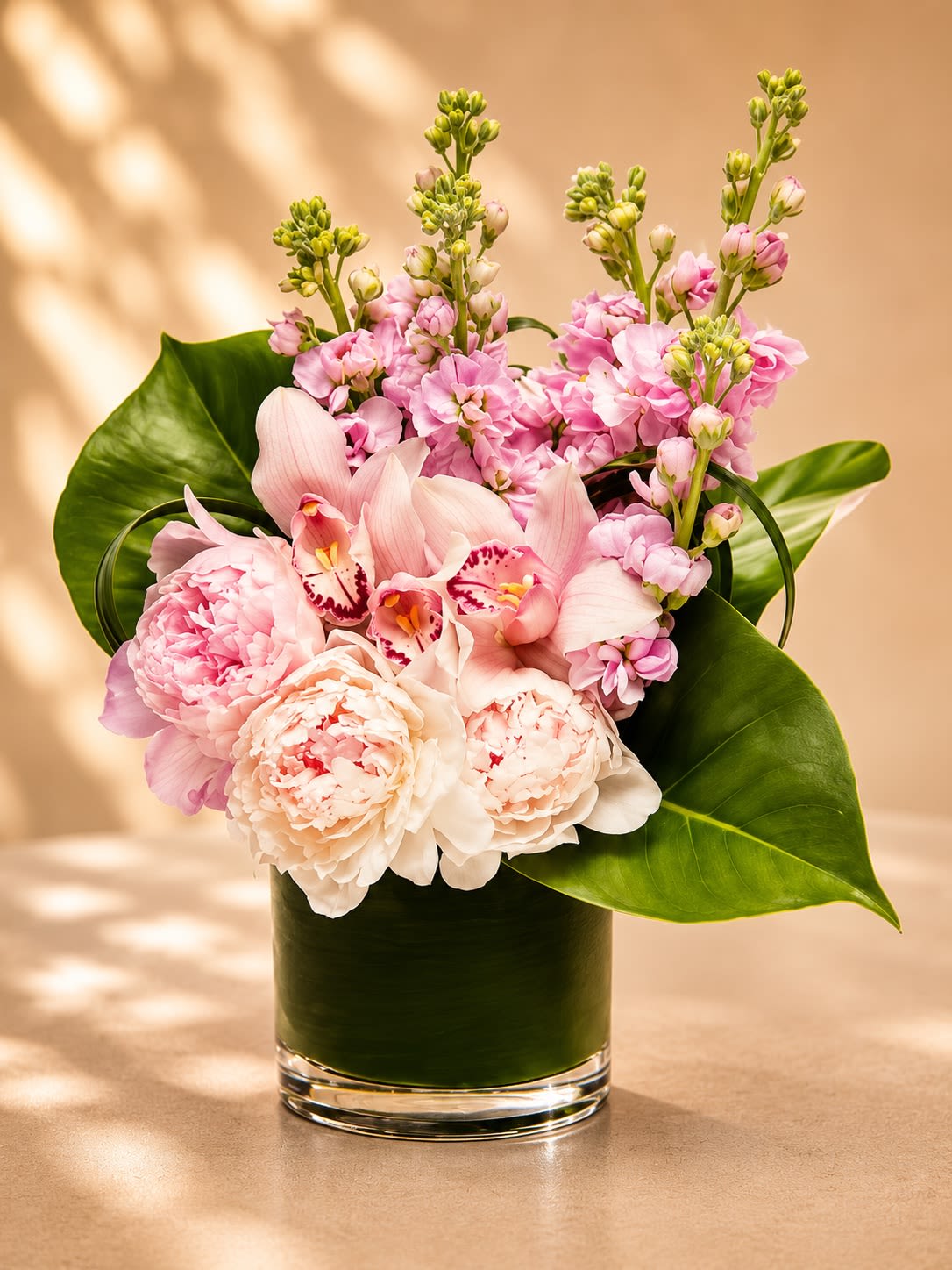 Pink Velvet Bloom - A luxurious garden-inspired arrangement featuring soft blush peonies, delicate pink cymbidium orchids, fragrant stock flowers, and dramatic tropical foliage arranged in a modern leaf-lined glass vase. Pink Velvet Bloom blends romantic textures with contemporary elegance, creating a floral design that feels graceful, feminine, and effortlessly upscale.  Peonies symbolize romance, prosperity, and beauty, while pink orchids represent admiration, refinement, and joy. The fragrant stock flowers add softness and movement, while sculptural tropical leaves bring a bold designer finish that elevates the entire arrangement. Perfect for anniversaries, birthdays, baby showers, luxury gifting, congratulations, or simply making someone feel deeply special.  Due to seasonal and market availability, peonies may occasionally be substituted with premium garden roses or similar luxury blooms while preserving the arrangement’s signature romantic blush palette and elegant modern style. Vase style and tropical foliage may vary slightly depending on availability while maintaining the same upscale presentation.
