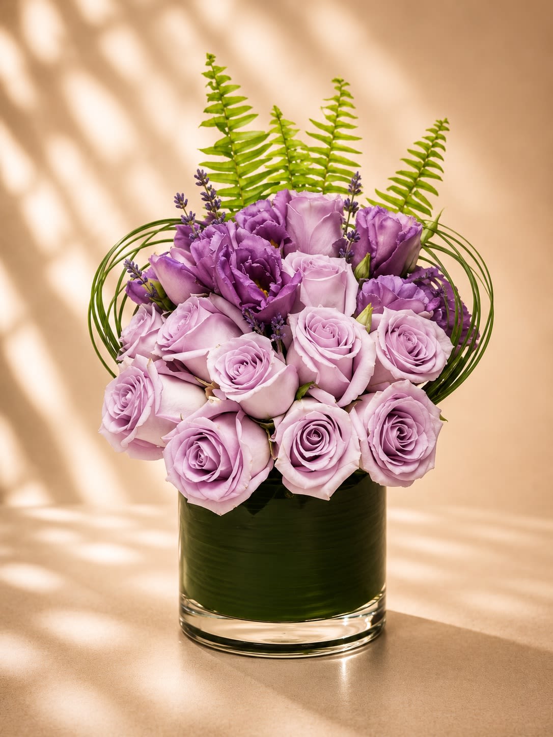 Lavender Amour - Soft, romantic, and effortlessly elegant, Lavender Amour features luxurious lavender roses paired with rich violet lisianthus, delicate lavender accents, sculptural greenery, and modern tropical foliage in a sleek leaf-lined glass vase.  This arrangement embodies sophistication and serenity with its dreamy monochromatic palette. Lavender roses symbolize enchantment, admiration, grace, and elegance, while lisianthus adds a refined garden-inspired softness that feels both modern and timeless.  Designed with elevated textures and artistic movement, Lavender Amour is perfect for anniversaries, birthdays, sympathy gestures, luxury gifting, romantic occasions, or simply bringing calming beauty into any space.  The airy fern accents and contemporary looping greens create a high-end couture floral look that feels fresh, feminine, and upscale — ideal for customers seeking a statement piece with soft luxury energy.