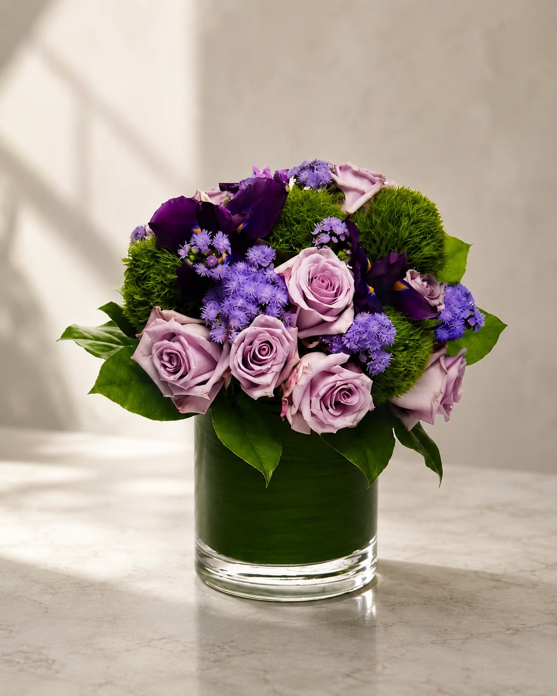 Violet Enchantment Garden - Elegant, artistic, and rich with texture, Violet Enchantment Garden blends soft lavender roses, deep violet calla lilies, vibrant green dianthus, and delicate lavender asters in a luxurious modern leaf-lined vase.  This arrangement captures a sophisticated jewel-toned palette inspired by twilight gardens and couture floral design. Lavender roses symbolize grace, admiration, and enchantment, while the dramatic purple calla lilies add depth, mystery, and modern elegance.  The lush green dianthus creates striking texture and contrast, giving the arrangement a fresh sculptural appearance that feels both contemporary and abundant. Delicate lavender asters soften the composition with airy romantic detail.  Perfect for birthdays, sympathy arrangements, luxury gifting, anniversaries, corporate floral décor, or customers looking for a refined and unique color palette.