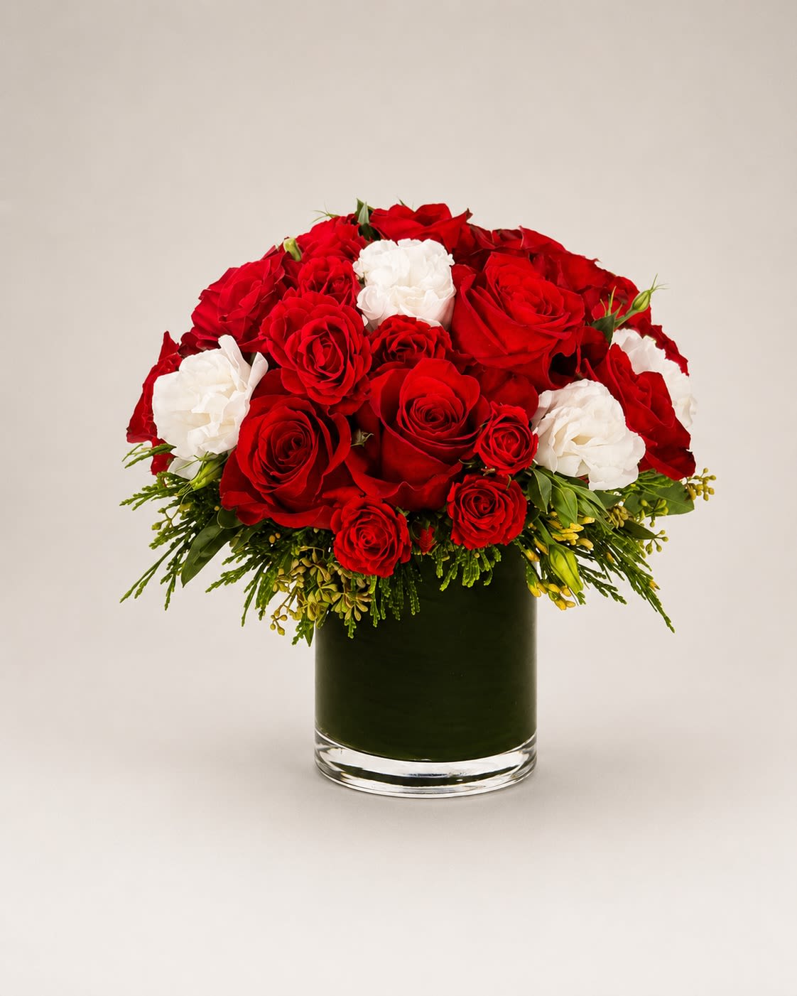 Ruby Ivory Romance - Elegant and timeless, Ruby Ivory Romance is a luxurious floral composition that blends passionate crimson roses with delicate ivory blooms for a classic yet elevated romantic aesthetic.  Designed in a sleek leaf-lined glass cylinder vase, this arrangement features premium red roses, lush spray roses, soft white lisianthus, and refined seasonal greenery that adds texture and movement. The contrast between deep ruby tones and soft ivory petals creates a balanced design that feels both bold and graceful.  This arrangement was inspired by modern luxury romance — sophisticated enough for upscale gifting while still warm, emotional, and inviting. Perfect for anniversaries, romantic gestures, weddings, engagements, holiday gifting, or elegant home décor.  The rich red roses symbolize deep love and devotion, while the ivory blooms represent sincerity, elegance, and everlasting connection.