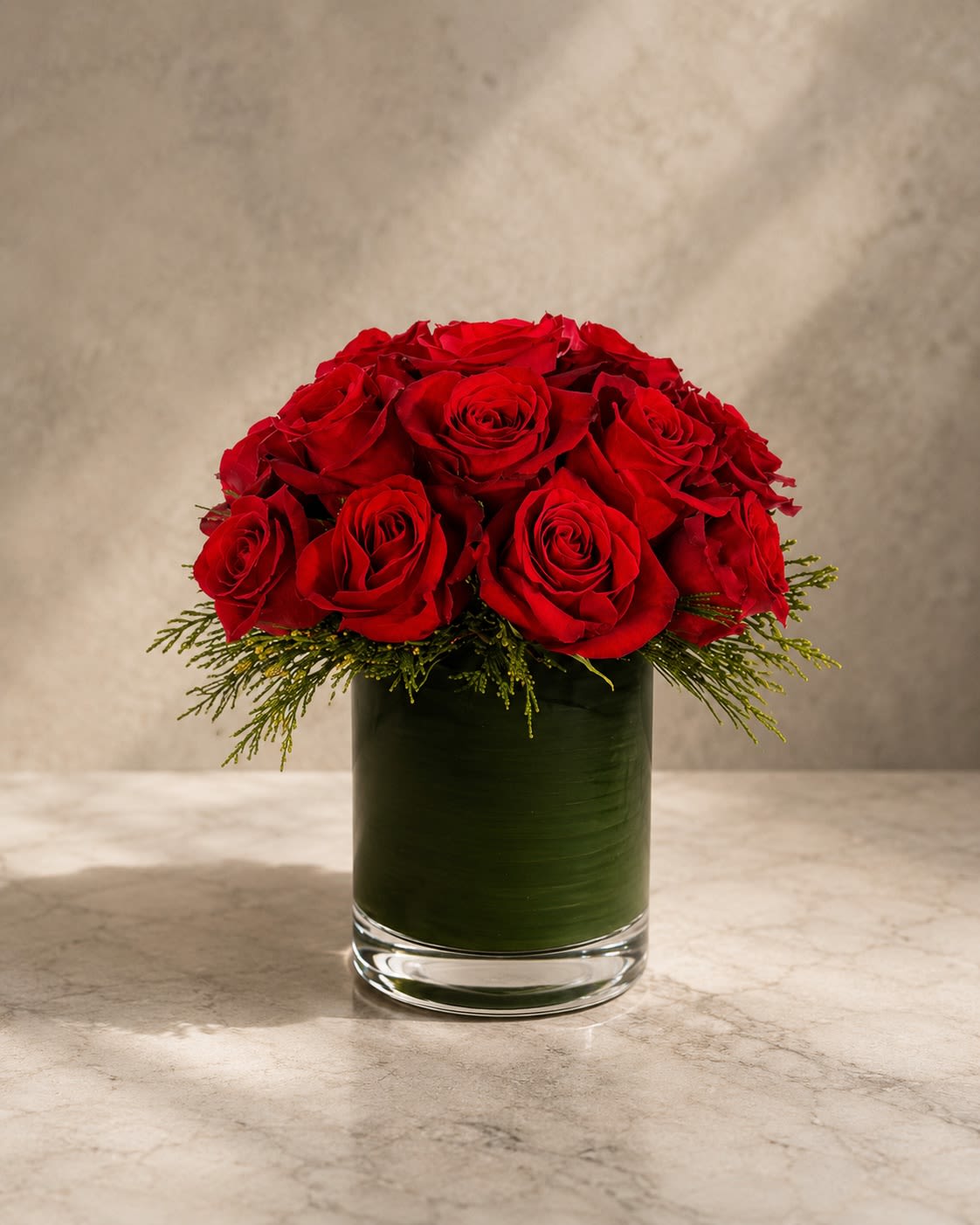 Scarlet Reverence - Classic, luxurious, and endlessly romantic, Scarlet Reverence is a timeless arrangement composed of premium long-stem red roses artfully gathered in a modern leaf-lined glass cylinder vase.  Designed with rich crimson roses and elegant evergreen accents, this arrangement embraces simplicity in its most sophisticated form. The rounded dome silhouette creates a refined, couture-inspired presentation that feels polished, graceful, and deeply romantic.  Each velvety red rose symbolizes passionate love, admiration, and devotion, making this arrangement perfect for anniversaries, Valentine’s Day, romantic celebrations, luxury gifting, or meaningful grand gestures.  Its clean monochromatic palette and minimalist styling give it the elevated feel of a luxury floral boutique centerpiece — timeless enough to never go out of style.