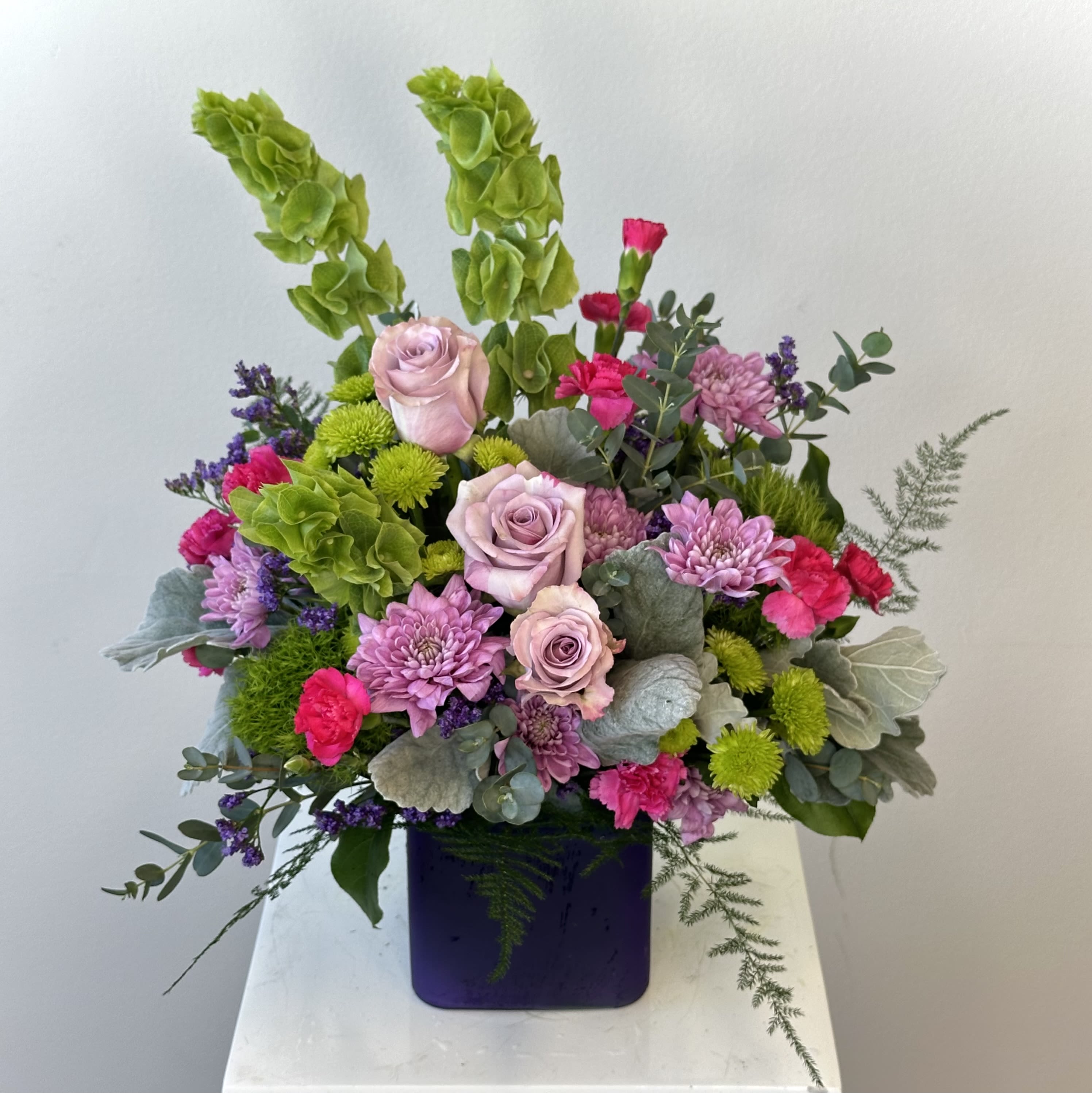 Purple Serenity Bouquet - Are you looking for a bouquet that makes a statement yet harmonizes into the interior of your home perfectly? Well, look no further than the Purple Serenity Bouquet! The lavender-colored roses paired with maroon blooms that are balanced out with the greens are all tied in together with an amethyst-colored cube - it's the perfect addition to your home as well as the perfect touching gift to a friend!   This bouquet features gorgeous pinky-lavender colored roses, bright red carnations, and cute pink chrysanthemums that are truly balanced out with the greenery.  Approximately: 16 1/4" W x 14 1/2" H  Orientation: All-Around