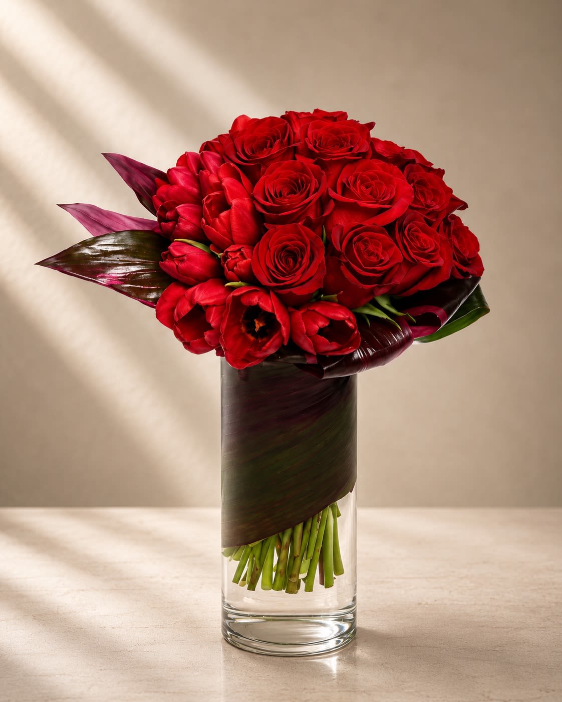 Rouge Eternal - Bold, romantic, and undeniably couture, Rouge Éternelle is a luxurious monochromatic floral design created to make a dramatic statement. Rich crimson roses and deep red tulips are artfully gathered into a lush modern silhouette, wrapped with glossy burgundy foliage inside a sleek clear glass vase for a refined editorial finish.  This arrangement embodies passion and sophistication through layered textures and tonal depth. The velvety roses symbolize timeless love and devotion, while the sculptural tulips add movement, softness, and contemporary elegance. The dark folded foliage creates a high-fashion presentation inspired by luxury floral ateliers and upscale hotel arrangements.  Perfect for anniversaries, romantic gestures, luxury gifting, Valentine’s Day, milestone celebrations, or elegant modern interiors, this arrangement is designed for clients who appreciate striking floral artistry with a couture aesthetic.