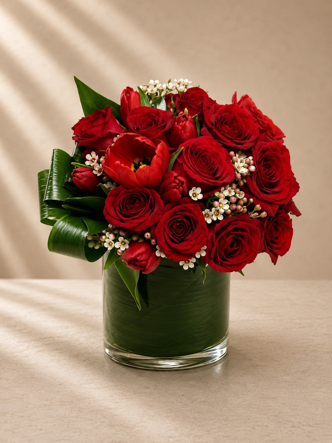 Velvet Rouge Garden - Romantic, lush, and exquisitely refined, Velvet Rouge Garden is a couture-inspired floral arrangement designed to capture timeless elegance with a modern luxury touch. Featuring premium red roses and velvety crimson tulips accented by delicate wax flowers and sculpted tropical foliage, this arrangement delivers richness, texture, and sophistication in every detail.  The layered red blooms create depth and passion, while the tiny white wax flowers soften the design with airy elegance and contrast. Glossy folded greenery adds an elevated architectural finish, giving the arrangement a polished high-end presentation worthy of luxury boutiques and upscale interiors.  Designed in a sleek leaf-lined glass cylinder vase, this arrangement feels both romantic and contemporary — perfect for meaningful gifting and statement décor.