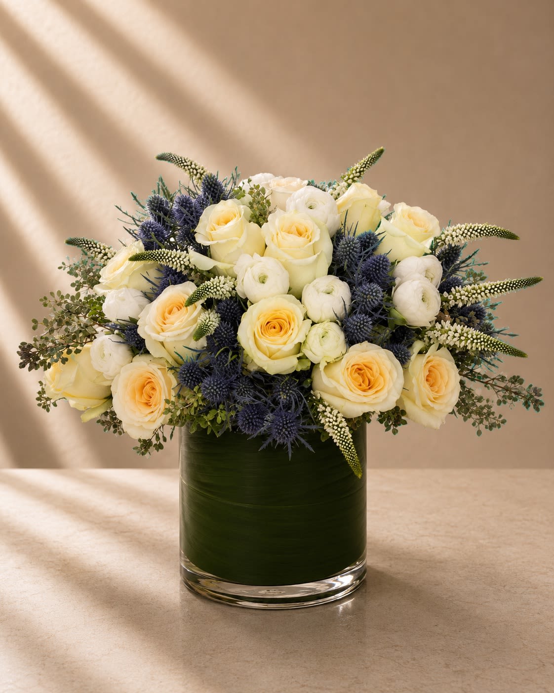 Ivory &amp; Indigo Luxe - Elegant, refined, and artfully textured, Ivory &amp; Indigo Luxe is a sophisticated floral arrangement that blends soft romance with modern botanical drama. Featuring creamy ivory roses and delicate white ranunculus accented with striking blue eryngium thistle and graceful veronica blooms, this design creates a luxurious balance of softness and bold texture.  The warm ivory roses bring timeless elegance and serenity, while the indigo-blue thistle adds depth, movement, and an unexpected couture edge. Airy greenery and elongated floral accents give the arrangement an organic garden-inspired silhouette while maintaining a polished upscale aesthetic.  Designed in a sleek leaf-lined glass cylinder vase, this arrangement feels fresh, elevated, and effortlessly luxurious — perfect for clients seeking something classic with a modern artistic twist.