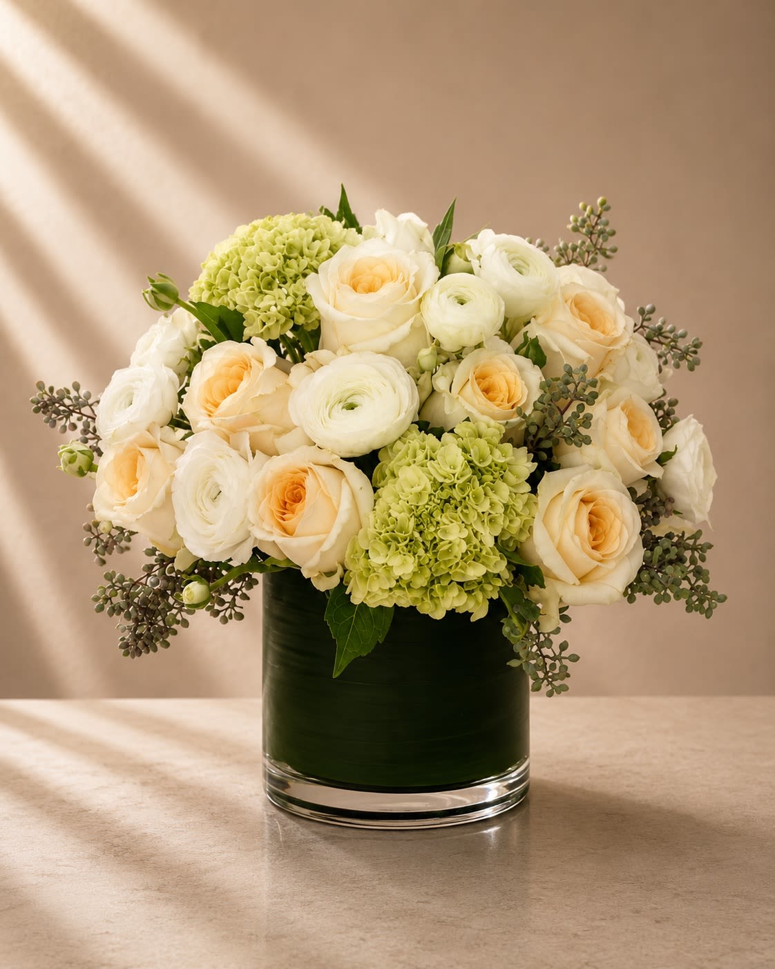 Ivory Garden Whisper - Soft, airy, and beautifully refined, Ivory Garden Whisper is a timeless luxury arrangement inspired by elegant European garden florals. Featuring creamy ivory roses, delicate white ranunculus, fresh green hydrangeas, and soft eucalyptus textures, this design radiates sophistication with a calm romantic presence.  The blend of lush blooms and layered textures creates a full, graceful silhouette while maintaining a clean modern aesthetic. Ivory roses bring warmth and elegance, ranunculus add delicate couture softness, and green hydrangeas introduce freshness and balance. Seeded eucalyptus accents complete the arrangement with organic movement and upscale botanical charm.  Designed in a sleek leaf-lined glass cylinder vase, this arrangement feels luxurious yet effortless — ideal for clients who love understated elegance and refined floral artistry.