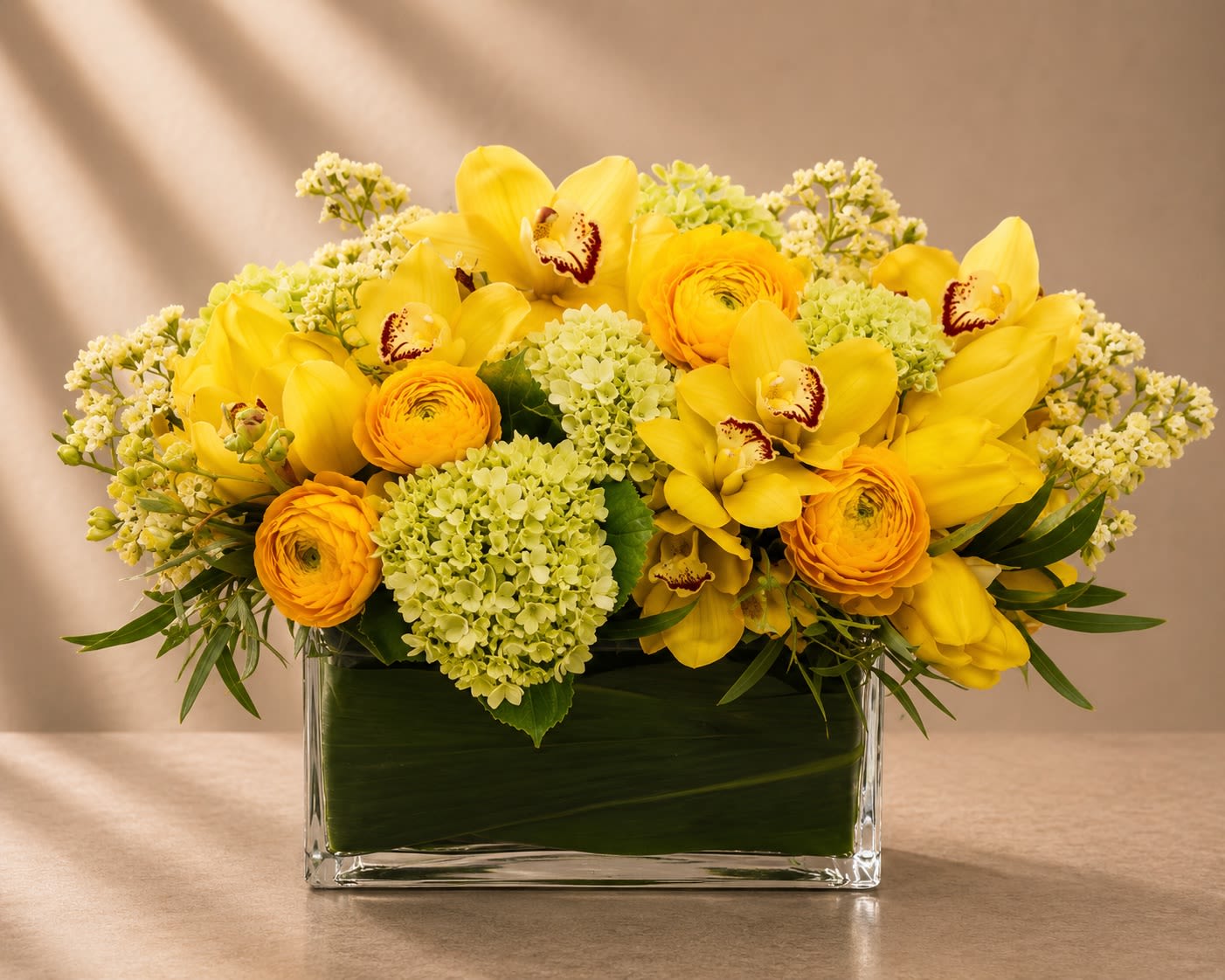 Soleil Jardin - Bright, joyful, and unmistakably luxurious, Soleil Jardin is a radiant garden-style arrangement featuring vibrant yellow tulips, golden ranunculus, cymbidium orchids, hydrangea, and delicate seasonal florals artfully designed in a modern glass rectangle vase.  Inspired by the warmth of a sunlit European garden, this arrangement blends soft texture with bold color to create a fresh, upscale statement piece. The layered yellow tones symbolize happiness and abundance, while the orchids and ranunculus add an elegant couture feel.  Its lush, sprawling composition gives the arrangement a premium designer aesthetic that feels both sophisticated and naturally effortless.
