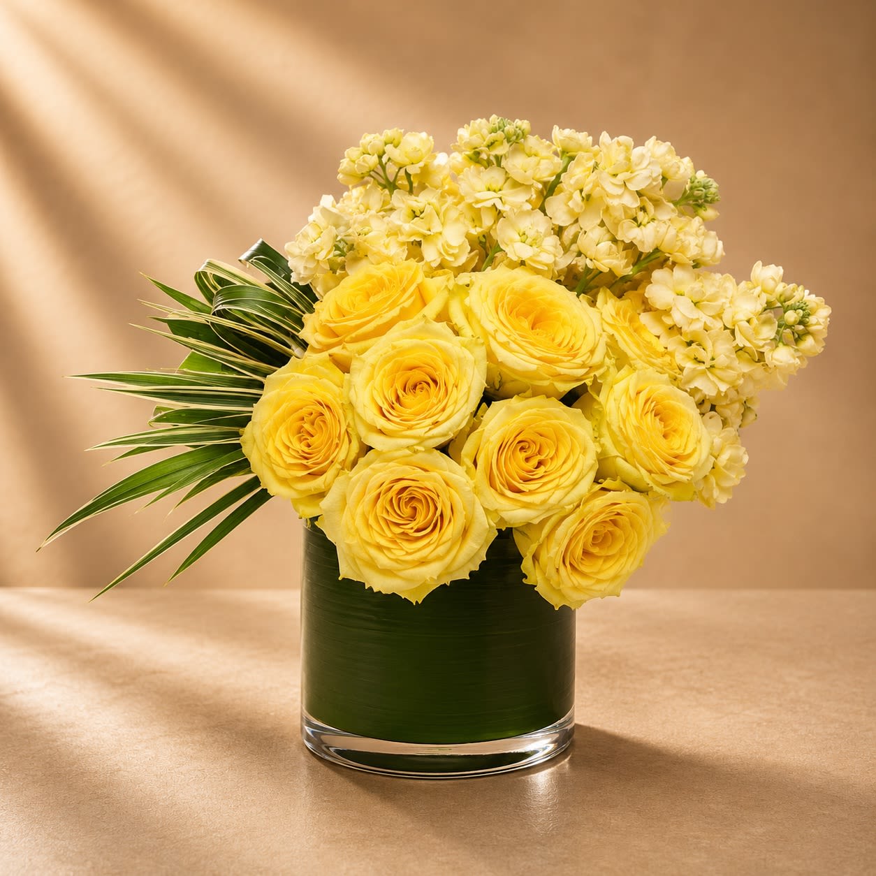 Maison Solara - Inspired by golden sunlight and refined botanical luxury, Maison Solara is a radiant floral arrangement featuring buttery yellow roses, delicate cream stock blossoms, and sculpted tropical greenery arranged in a sophisticated contemporary style.  Warm tones and soft textures create an atmosphere of elegance and serenity, while the architectural foliage gives the design a couture-inspired finish. Bright yet graceful, this arrangement embodies modern luxury with a luminous garden aesthetic.