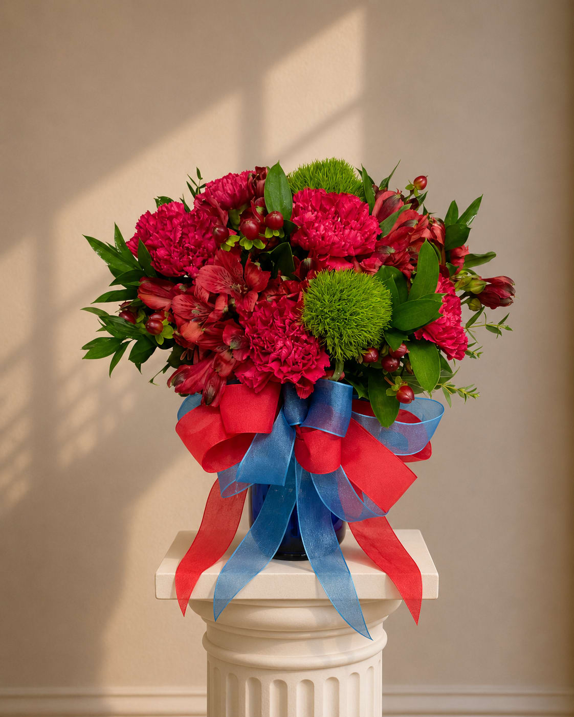 Bold &amp; Beautiful - A lively cluster of bright pink carnations and vivid red alstroemeria arranged with round green accent blooms creates an immediate burst of color-perfect for a birthday bouquet or a congratulations arrangement that needs to feel joyful and festive. The bold pink and red palette reads celebratory, while the pops of green add texture so the person you're sending it to feels noticed and genuinely cheered.  Flowers are gathered tightly in a dark glass vase and finished with oversized red and blue ribbons for a playful, party-ready look. Carnations give soft, ruffled texture; alstroemeria adds delicate stripes and longevity; the round green accents bring a fresh contrast that keeps the design energetic without feeling fussy. This arrangement arrives ready to display in the included glass vase; change the water every few days and trim stems for longest life.