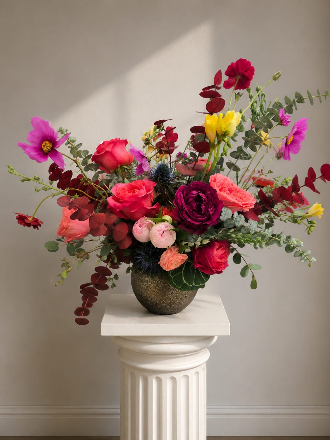 Just B Kind - Bold, garden-fresh color greets the recipient with this lively flower arrangement-perfect as a birthday bouquet, anniversary surprise, or a bright thank-you gesture. Loose, airy stems in vivid pinks, reds, yellows, purples, and mixed tones create a playful yet romantic statement that makes the person you're thinking of feel celebrated and genuinely seen.  A mix of anemones, ranunculus, roses, daisies, carnations and other seasonal blooms are gathered in a dark ceramic vase set on a round wooden base. Layered, round blossoms contrast with daisy-like faces and ruffled petals while eucalyptus-style foliage and trailing stems add natural movement and an asymmetrical, garden-inspired silhouette. The arrangement's size and fullness read as generous without being formal, bringing a contemporary, hand-picked look to any room.  Ideal for everyday gifting or marking special moments, this arrangement arrives ready to display in its ceramic vessel on the included wooden base. To keep blooms looking fresh, replace vase water every 2-3 days and trim stems at an angle if you transfer them-small care steps that help the bouquet stay vibrant through the week.