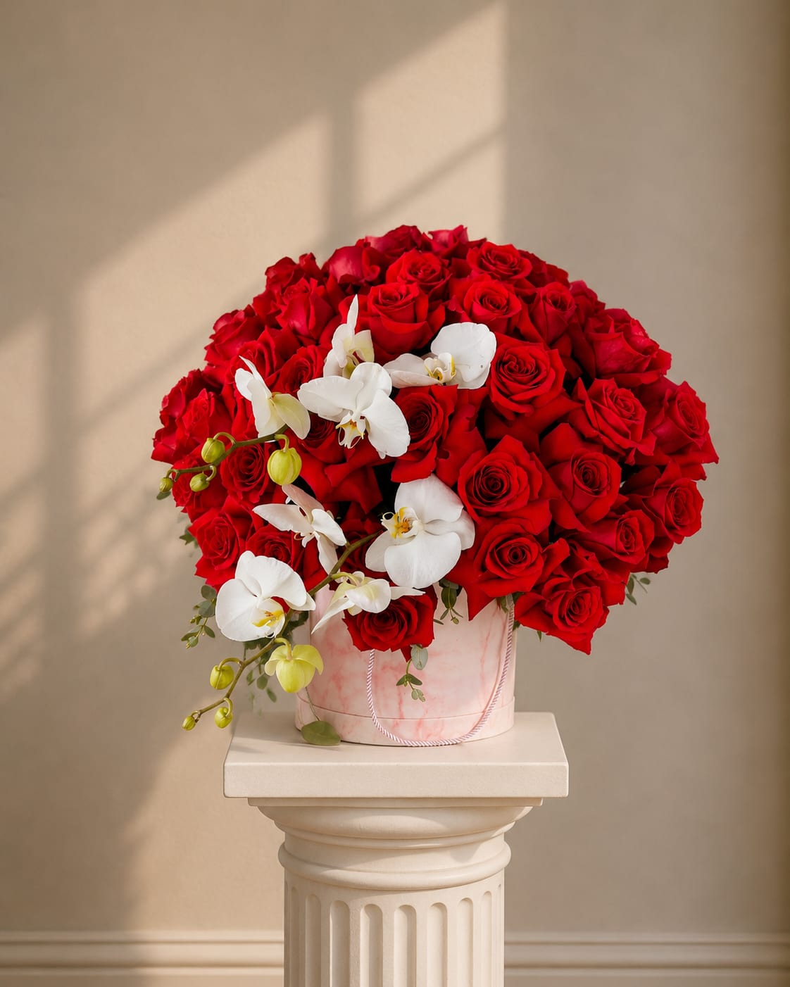 Triumphant - A lush dome of vivid red roses crowned with cascading white phalaenopsis orchids creates a romantic, dramatic statement - perfect as a birthday bouquet, anniversary roses gift, or a Valentine's Day arrangement that reads both bold and tender. Triumphant is designed to make the person you're thinking of feel deeply celebrated: the dense, velvety roses deliver passion while the orchids add a graceful, whispering contrast.  The arrangement is presented in a pale pink hatbox with a rope handle for an elegant, ready-to-gift finish. Flowers are tightly packed for a full, luxurious silhouette; red roses dominate the composition while white Phalaenopsis spill gently over the side, with hints of soft pink from the container tying the palette together. Ideal for romantic occasions or upscale celebrations, this piece arrives polished and ready to place on a table or bedside - the kind of bouquet that makes someone feel truly seen.