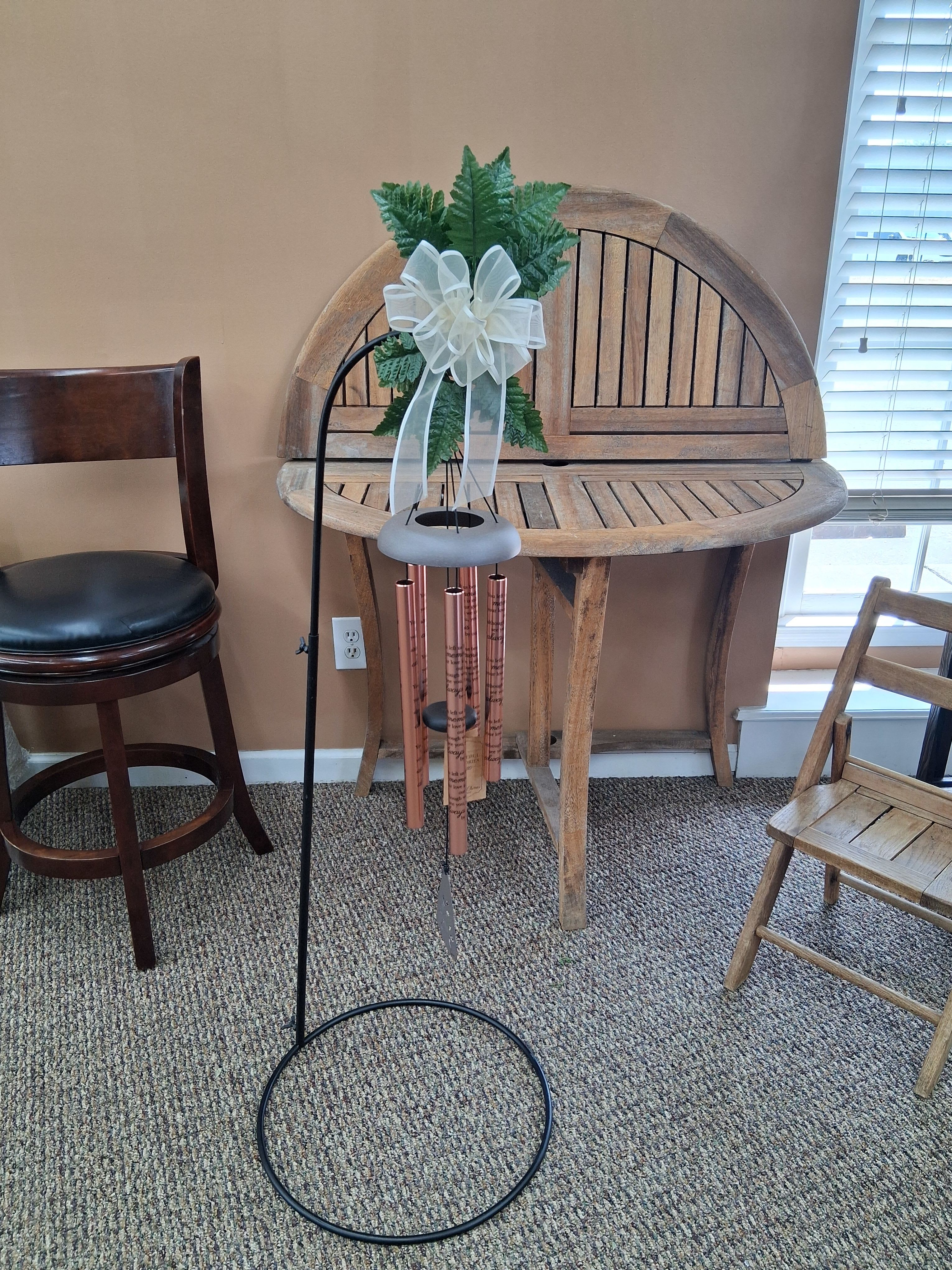 Beautiful Memories Wind Chime Rose Gold- Huntsville sympathy delivery - A rose-gold memorial wind chime that reads like a quiet tribute - perfect as a sympathy or funeral keepsake to honor someone you miss. The hanging wind chime is displayed on a black metal stand and topped with a soft white ribbon bow and faux greenery, so it arrives ready to gift and display on a porch, garden, or memorial spot. The engraved tubes and attached "Beautiful Memories" tag make the sentiment clear without words being overly ornate.  The chime's copper/rose-gold tubes carry black remembrance text and produce a gentle, comforting sound; the light gray top and understated palette of white, green, pink and subtle orange accents keep the look rustic and homey. Included hard goods are the wind chime, black metal stand, white ribbon bow, decorative greenery, and a paper hanging tag for the message.  This is the kind of keepsake that helps someone feel remembered long after the service - a thoughtful gift for family, a bereaved friend, or to place at a gravesite or memorial garden. It ships ready to hang on the included stand; minimal care is needed beyond occasional wiping to preserve the finish.