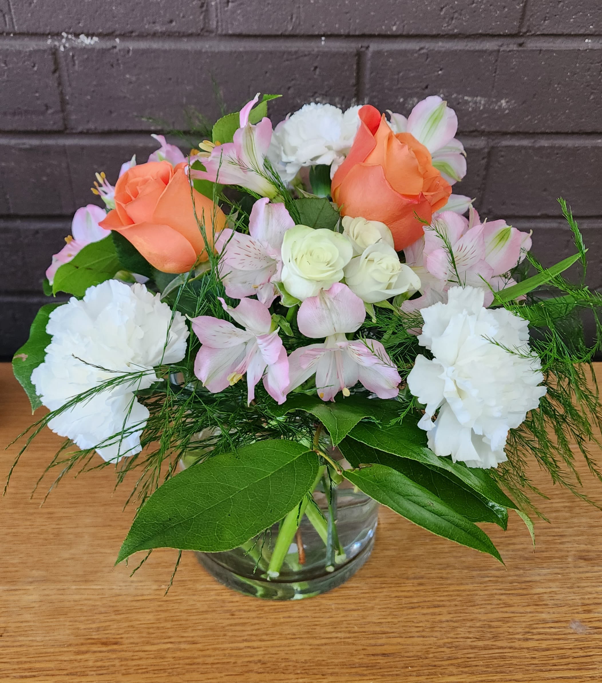 Summer Peach - Peach roses take center stage in this Summer Peach flower arrangement, their warm glow balanced by white roses, pale pink alstroemeria, and crisp white carnations-an ideal birthday bouquet, thank-you gift, or spring celebration arrangement. The soft, fresh palette and garden-style silhouette create a welcoming, uplifting impression that will make the person you're thinking of feel noticed and genuinely celebrated.  Arranged in a clear glass vase, the mix of textures-from the velvety rose petals to the delicate alstroemeria and ruffled carnations-is finished with airy filler and broad green leaves for fullness. The compact, hand-tied shape sits beautifully on a dining table or desk; its colors read as both elegant and cheerful, perfect for sending warm wishes without being over-the-top.  This everyday arrangement is designed to arrive ready-to-display in water-filled vase packaging so your recipient can enjoy instant beauty and a light, fresh fragrance. For care, keep the vase topped up with fresh water, trim stems slightly every few days, and keep away from direct heat to extend the life of the blooms.