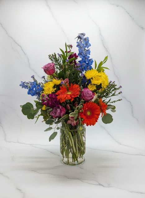 Color Me Happy - A vibrant and cheerful arrangement bursting with bold color and fresh seasonal blooms. Featuring bright gerbera daisies, roses, delphinium, chrysanthemums, and accent flowers arranged in a clear glass vase, this design is perfect for birthdays, celebrations, get well wishes, congratulations, or simply brightening someone's day. Its lively mix of colors creates an uplifting display that brings joy to any room.