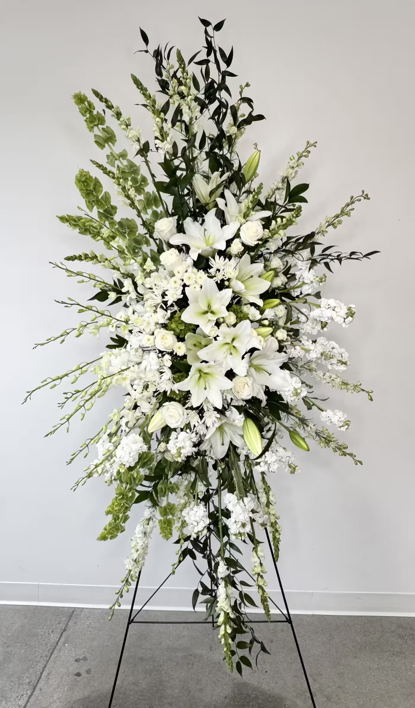Grand Farewell - Grand Farewell is an extraordinary sympathy spray designed to create a breathtaking and unforgettable tribute in honor of a loved one. Standing approximately 7ft tall and 3.5ft wide, this grand floral display offers an elegant expression of peace, remembrance, and heartfelt sympathy for funerals, memorial services, celebrations of life, and graveside ceremonies. Featuring an all-white palette of premium fresh flowers, Grand Farewell combines timeless beauty with graceful sophistication to provide comfort and honor during life’s most difficult moments.  This luxurious arrangement includes fragrant white lilies, elegant white stock, towering white snapdragons, lush white roses, striking Bells of Ireland, and delicate dendrobium orchid buds. The crisp white blooms symbolize purity, peace, reverence, and eternal love, while the vibrant green Bells of Ireland add height, texture, and a symbol of hope and good fortune. Together, these flowers create a dramatic and serene presentation that beautifully reflects a life remembered with love and admiration.  Displayed on a standing easel, Grand Farewell makes a stunning focal piece suitable for funeral homes, churches, memorial chapels, and large services. Each arrangement is handcrafted by our florists using premium flowers to ensure every tribute is both visually impactful and deeply meaningful. Grand Farewell serves as a powerful expression of sympathy, respect, and everlasting remembrance for someone who will always be cherished.