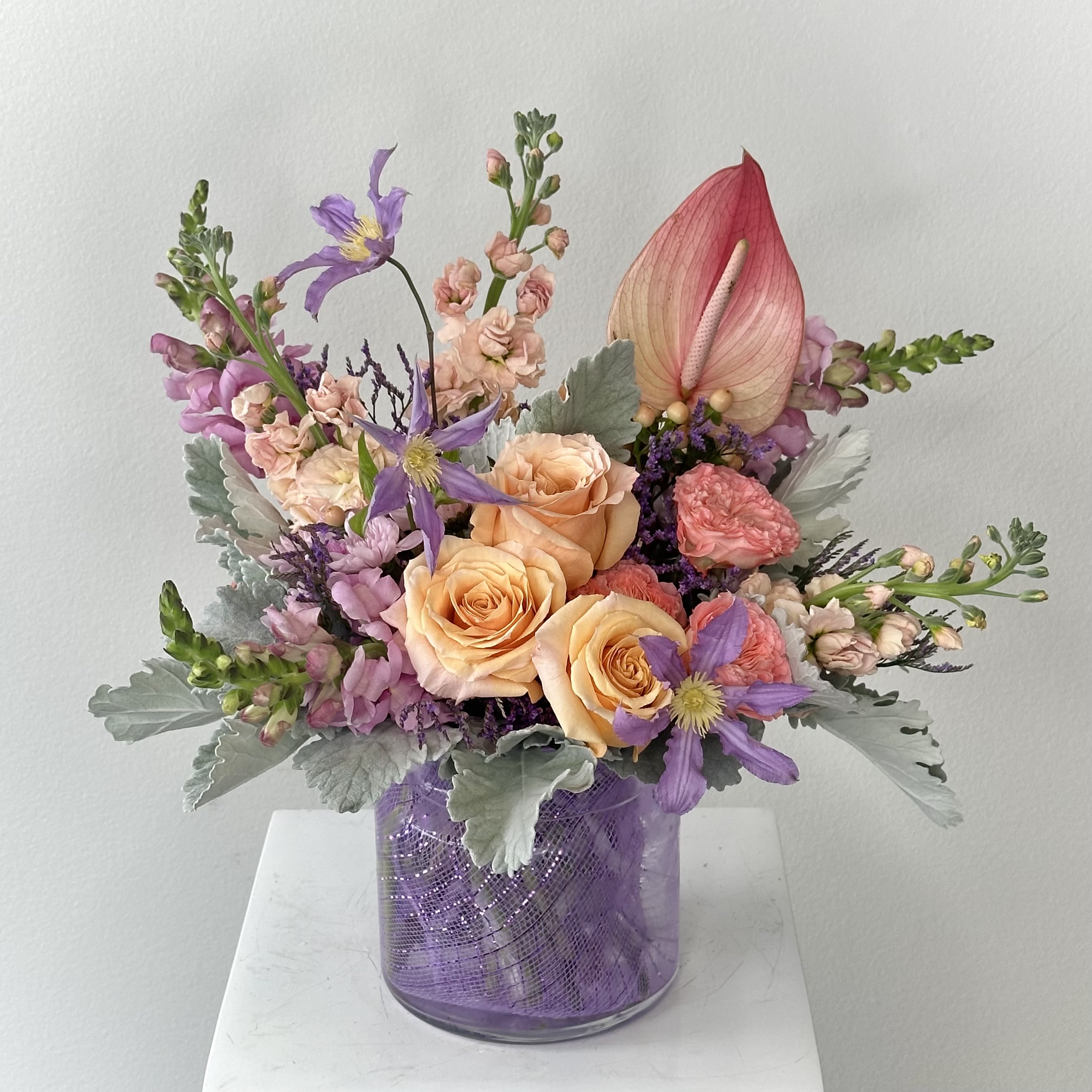 Fantasia Bouquet - Soft, romantic, and full of delicate charm, this pastel flower arrangement is designed to bring warmth and elegance to any occasion. Featuring a dreamy blend of peach roses, peach stock, lavender snapdragons, pink garden spray roses, lavender clematis, lavender cushion mums, and a striking pink anthurium, this arrangement showcases a beautiful harmony of soft tones and layered textures. Accented with silvery dusty miller and purple limonium, the design is balanced with gentle greenery that enhances the overall pastel palette.  Presented in a vase lined with lavender lace, this arrangement offers an extra touch of detail that perfectly complements the soft lavender hues throughout the design. The lace lining adds a subtle, romantic finish, making the entire piece feel elevated and thoughtfully curated.  Ideal for birthdays, celebrations, baby showers, or simply brightening someone’s day, this arrangement captures the essence of spring and timeless beauty. The combination of soft peach, blush pink, and lavender tones creates a soothing yet visually captivating display that works beautifully in any setting.  Whether you're sending a heartfelt gift or decorating a special space, this pastel floral arrangement delivers a graceful and elegant statement with its refined color palette and lush, garden-inspired design.