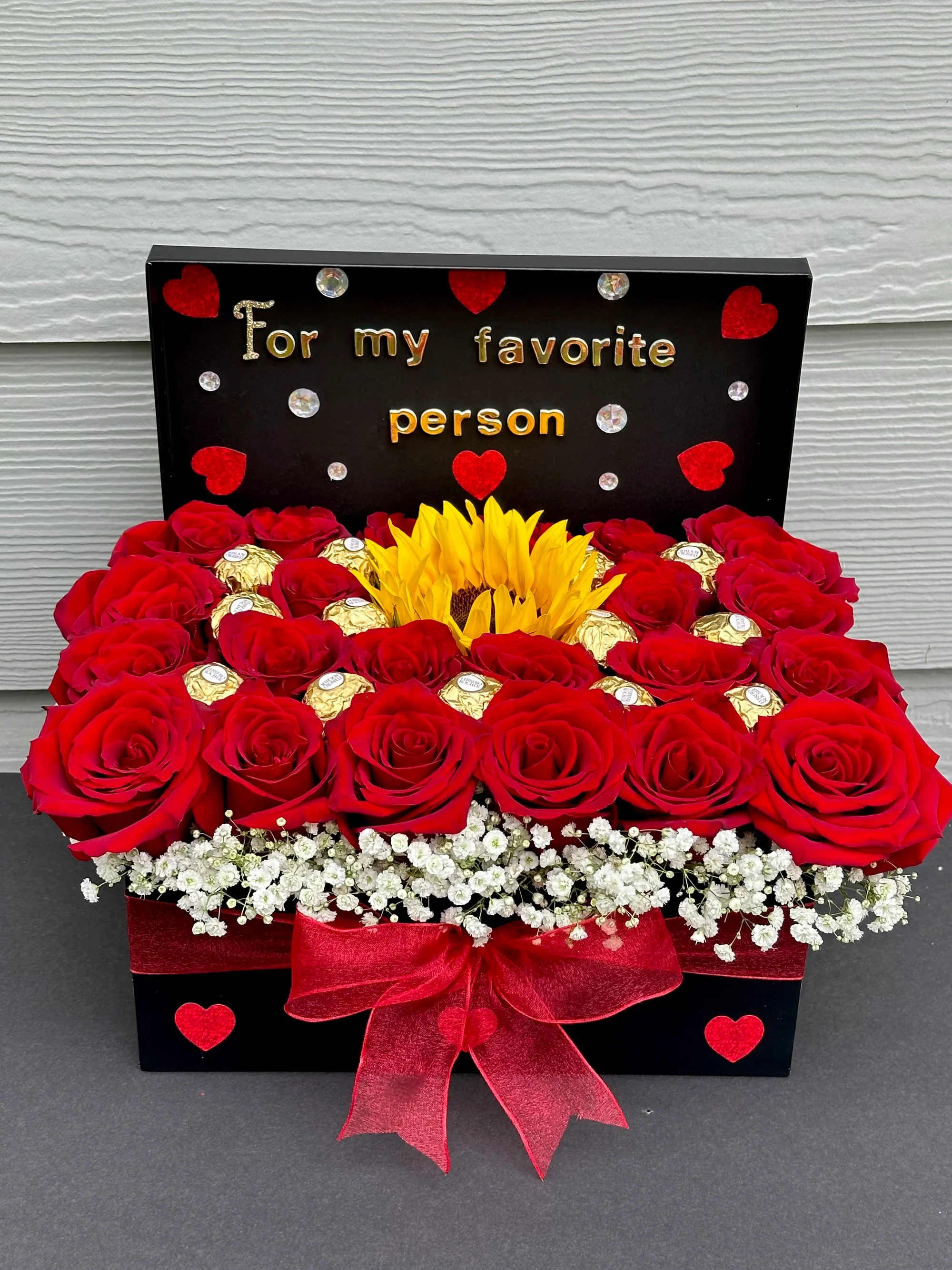 Cajita de rosas - Un detalle elegante y lleno de significado para esa persona que ocupa un lugar especial en tu corazón. Este hermoso arreglo combina la intensidad de las rosas rojas con la alegría de un girasol central y el toque dulce de chocolates premium.  Incluye:   Rosas rojas frescas de alta calidad  Girasol natural como punto central del diseño  Chocolates premium cuidadosamente integrados  Delicadas flores blancas decorativas (baby’s breath)  Moño elegante en tono rojo  Caja decorativa con mensaje “For my favorite person”  Perfecto para cumpleaños, aniversarios, San Valentín o simplemente para sorprender con amor y gratitud.   Diseño exclusivo y elaborado artesanalmente.  Disponible para entrega especial.