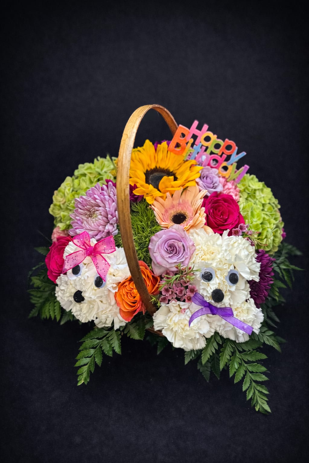 Trixi &amp; Tigger Dog Bouquet - Two playful dog figures made from tightly clustered white carnations steal the show in this whimsical flower arrangement - a cheerful gift-style bouquet perfect for birthdays, a "just because" surprise, or a lighthearted Father's Day nod. The floral pups wear googly eyes and sweet black pom-pom noses, one finished with a purple ribbon bow and the other with a black bow, so the person you're thinking of will smile the moment they see it.  Surrounding the dogs is a lively mix of pink and lavender roses, sunny yellow-orange gerbera daisies, colorful chrysanthemums, alstroemeria, and a few Asiatic lilies for height and fragrance. Everything is arranged in a compact wicker basket with a wooden handle, creating a finished, gift-ready presentation that sits nicely on a table or desk. Bright colors and varied textures give the arrangement a playful, celebratory feel ideal for thank-you gestures, congrats, or simply to let someone know you care.  This compact size makes it an easy pick-me-up for children or animal lovers and a charming alternative to a traditional vase bouquet. Order for same-day delivery when available to ensure your recipient gets that instant, joyful reaction.