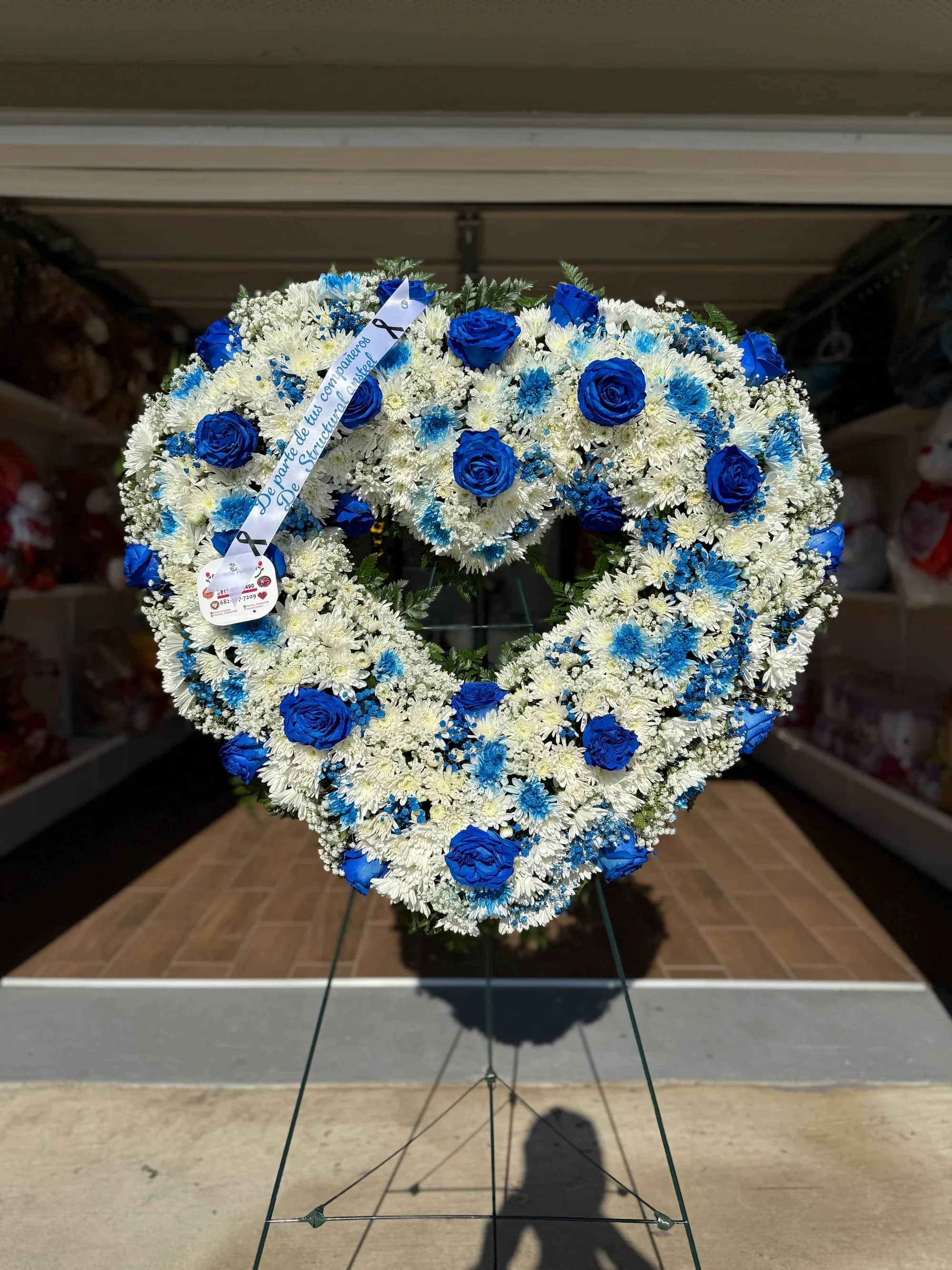 Corazon funeral  -  Corona Fúnebre en Forma de Corazón – Tonos Blanco y Azul   Honra la memoria de tu ser querido con esta elegante corona fúnebre en forma de corazón, diseñada en delicados tonos blanco y azul que simbolizan paz, esperanza y amor eterno.  Elaborada con una armoniosa combinación de rosas azules, crisantemos blancos y flores de complemento, esta pieza destaca por su diseño especial en forma de corazón, ideal para expresar un último adiós lleno de cariño y respeto.   Detalles del producto:  Diseño en forma de corazón Rosas azules Crisantemos blancos Flores de relleno delicadas Follaje verde natural Incluye tripié para exhibición Espacio para banda o mensaje personalizado   ️ Ideal para funerales, servicios memoriales o ceremonias de despedida.   Un arreglo sobrio y significativo para expresar condolencias con elegancia.