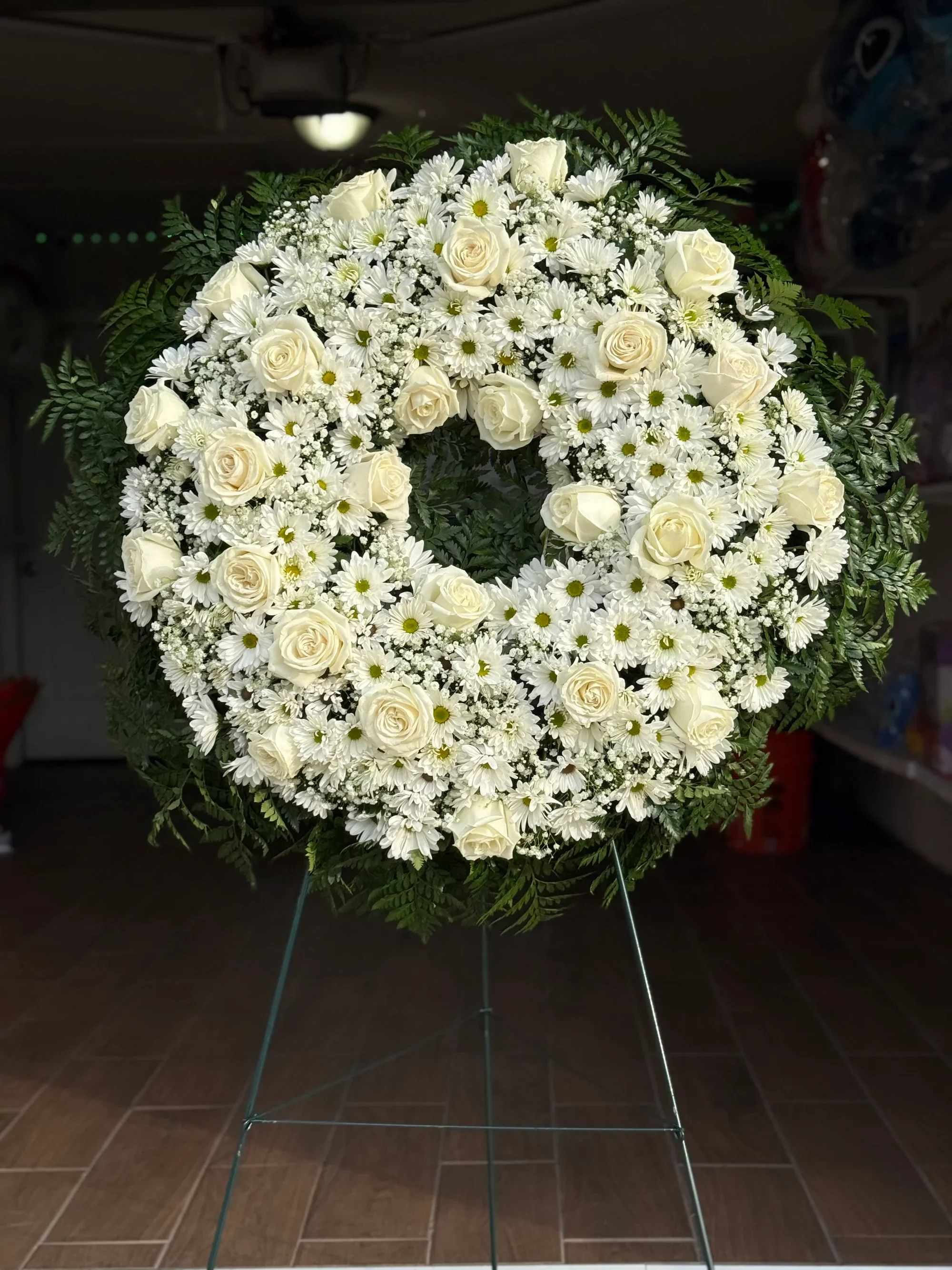 Corona 2 -  Corona Fúnebre Blanca “Paz Eterna” ️   Expresa tus condolencias con esta hermosa corona fúnebre en tonos blancos, símbolo de pureza, respeto y amor eterno. Su diseño circular representa la eternidad y el recuerdo que permanece para siempre en el corazón.  Elaborada con una delicada combinación de rosas blancas, margaritas, crisantemos y baby breath, complementadas con follaje verde natural que realza su elegancia y solemnidad.  Un arreglo clásico y distinguido, ideal para rendir homenaje con sensibilidad y buen gusto.    Detalles del producto:  Rosas blancas frescas Margaritas blancas Crisantemos blancos Flores de relleno (baby breath) Follaje verde natural Base metálica incluida    ️ Ideal para funerales, servicios memoriales o ceremonias de despedida.   Un tributo floral que transmite paz y acompañamiento en momentos difíciles.