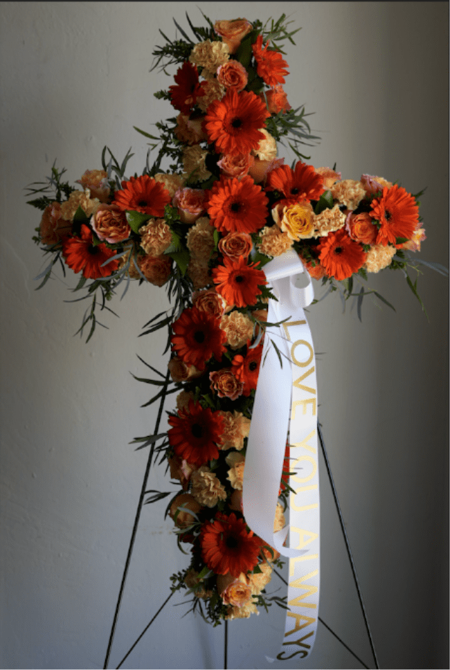 Cross Standing Spray  - A bold cross-shaped standing spray crafted for memorial and funeral services, covered in bright orange gerbera daisies, peach and yellow roses, and pale carnations to create a warm, reverent presentation.