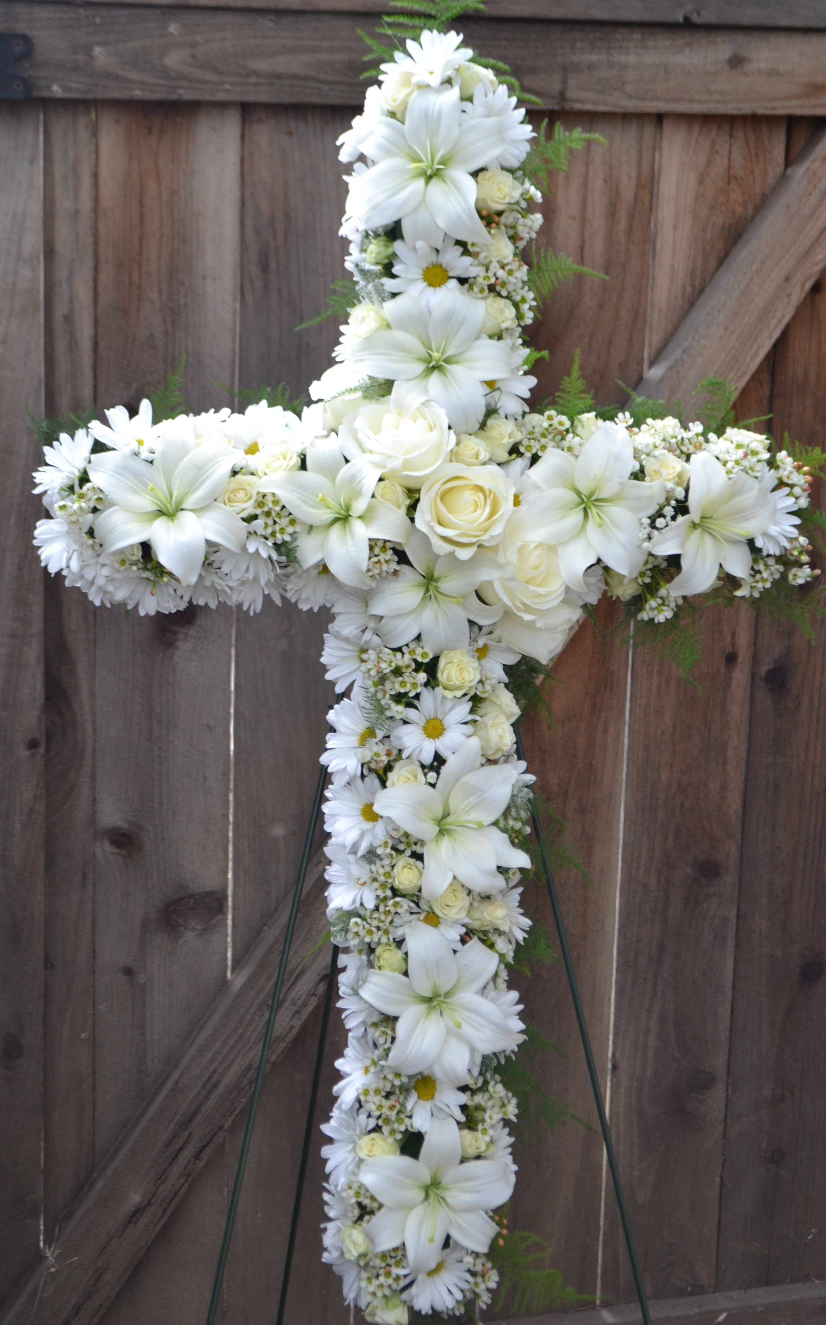 Memorial Cross - Memorial Cross is a timeless sympathy easel designed to honor the life and memory of a loved one with grace, faith, and beauty. This elegant standing cross arrangement features a lush combination of fresh lilies, roses, daisies, and chrysanthemums, thoughtfully arranged to create a meaningful tribute for funerals, memorial services, church ceremonies, and celebrations of life. Available in one color or multiple custom color combinations, the Memorial Cross can be designed to reflect personal preferences, favorite colors, religious significance, or a loved one’s unique spirit.  Lilies symbolize peace and the restored innocence of the soul, while roses represent love and remembrance. Daisies offer a gentle expression of purity and hope, and mums are traditionally associated with honor, sympathy, and lasting devotion. Together, these flowers create a beautiful and comforting display that serves as a touching expression of support and condolences during difficult times.  Displayed on a standing easel, the Memorial Cross creates an elegant presentation suitable for funeral homes, churches, graveside services, and memorial gatherings. Each arrangement is handcrafted by our florists using fresh, premium blooms to ensure every tribute is both meaningful and visually stunning.  Please allow at least one day’s notice when ordering the Memorial Cross, as custom color combinations and floral availability may require additional preparation time. Same-day orders are not guaranteed.