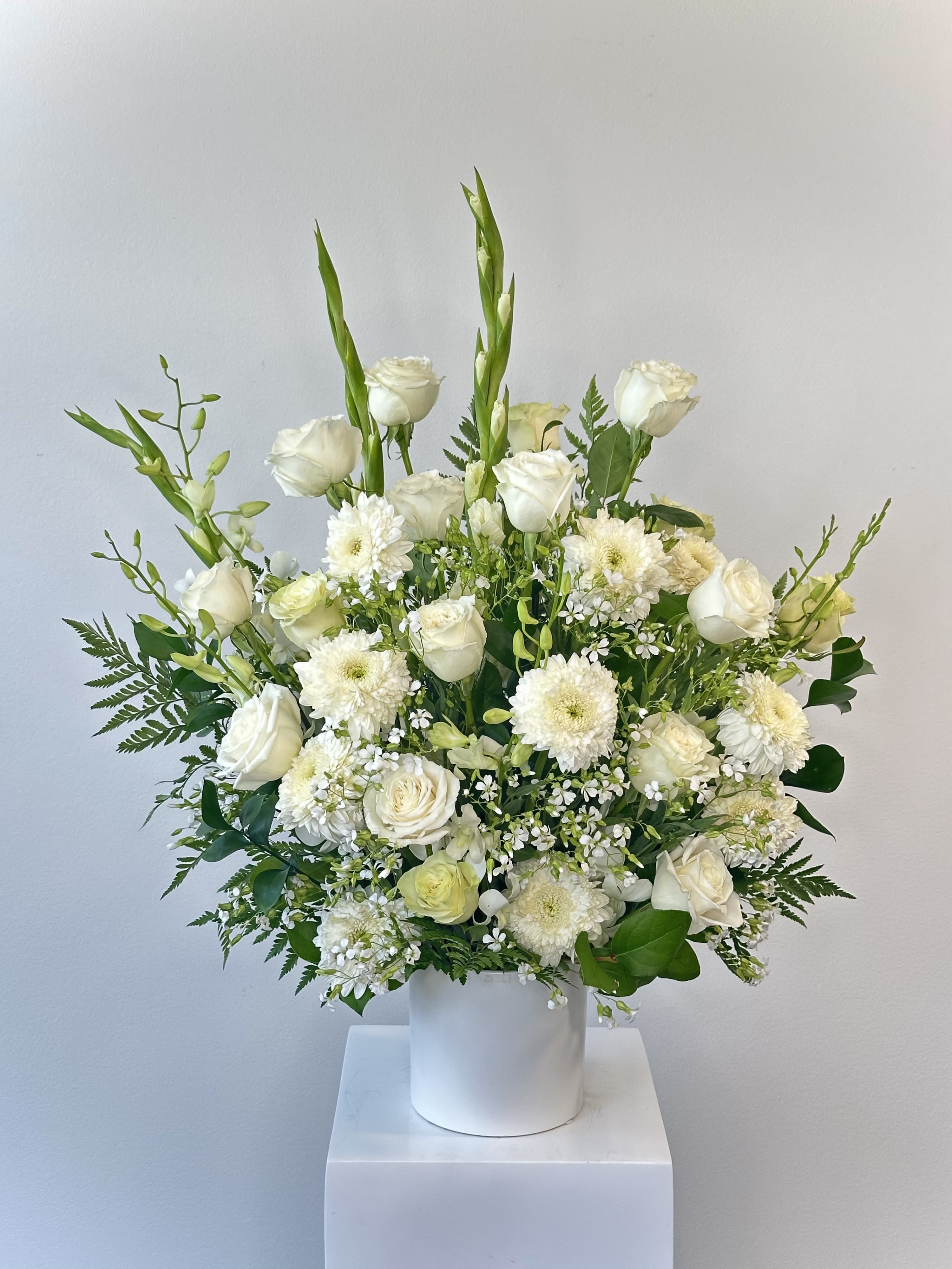 Venice - Bring timeless elegance and graceful beauty to the reception with the Venice flower arrangement, a stunning all-white design inspired by classic European charm. This tall, sophisticated arrangement features a beautiful blend of fresh white blooms carefully arranged to create height, texture, and a soft, luxurious look.   Elegant white gladiolas rise above the design, adding dramatic height and a graceful silhouette that makes the arrangement truly eye-catching. Classic white roses bring romantic beauty and smooth, layered petals, while full white mums add lush texture and volume throughout the arrangement. Delicate small white filler flowers weave between the larger blooms, creating a light, airy feel and enhancing the arrangement’s natural elegance. Fresh greenery adds depth and contrast, highlighting the purity of the white flowers while giving the design a balanced, garden-inspired finish.  Presented in a clean, modern white ceramic vase, the Venice arrangement has a refined look that complements both traditional and contemporary décor. Standing approximately 28 inches tall and 22 inches wide, this impressive arrangement makes a beautiful statement for sympathy occasions.  While the classic white version offers timeless sophistication, the Venice arrangement can also be custom created in a variety of color palettes to match your occasion, décor, or personal style, making it a versatile and memorable floral centerpiece.